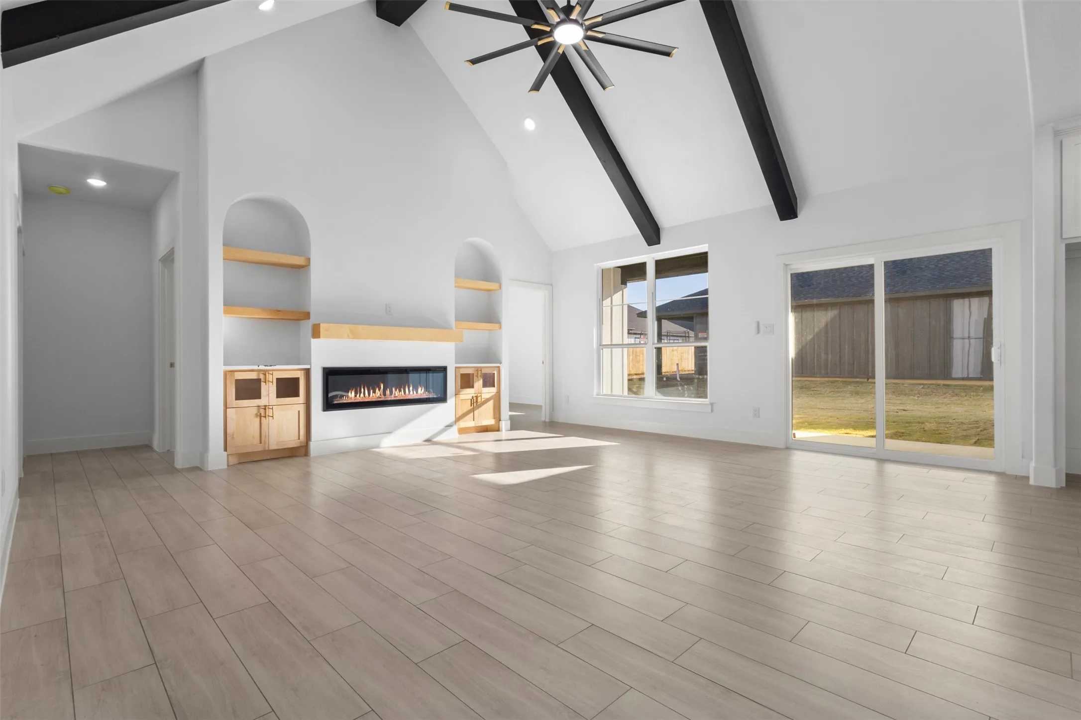 Vaulted ceiling with exposed beams, wood-finish flooring, built-in shelving with lower cabinetry, linear electric fireplace, and sliding glass door to exterior