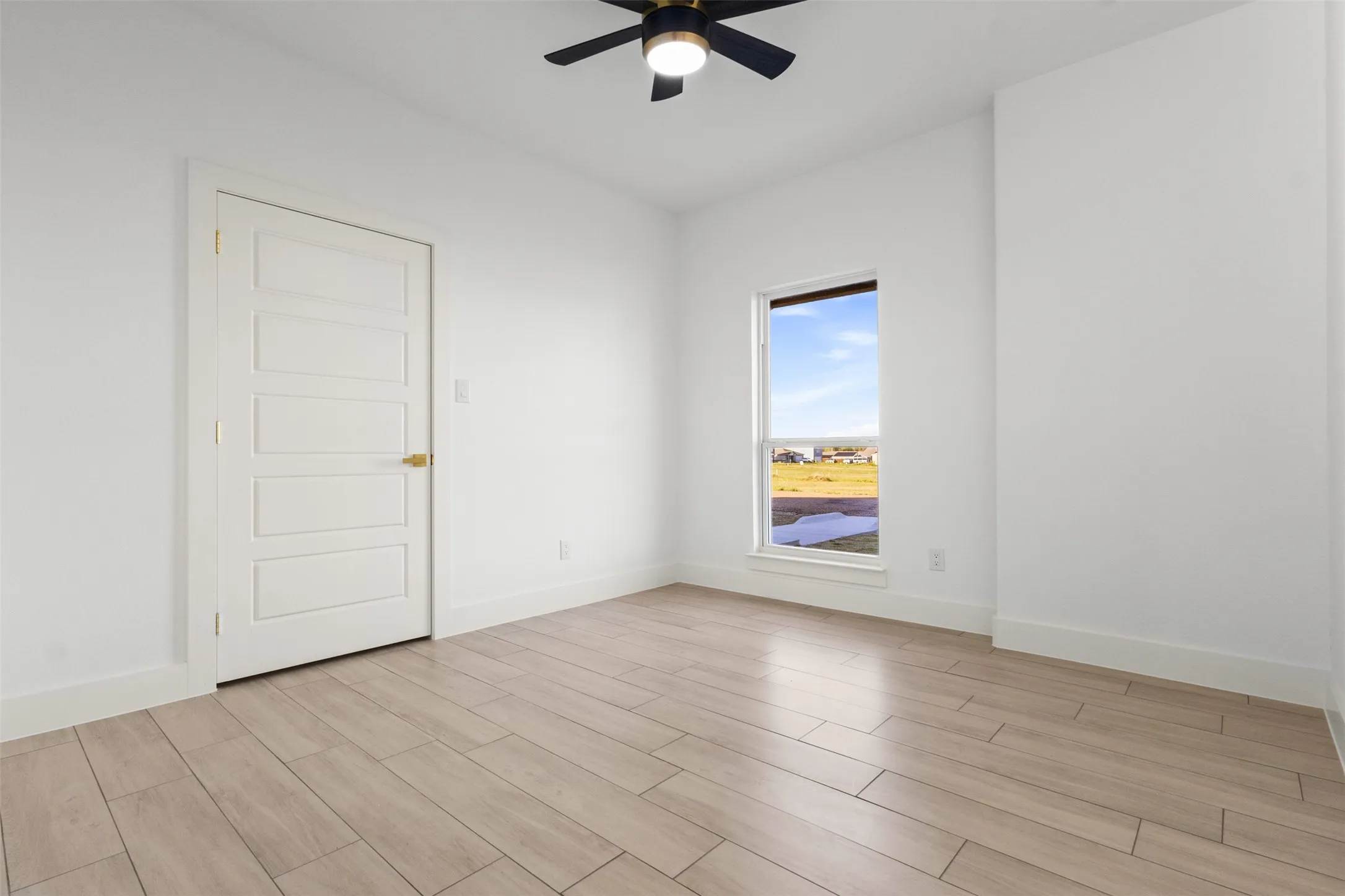 Wood-finish flooring, white walls, five-panel interior door with brass hardware, ceiling fan with integrated lighting, and a vertical window