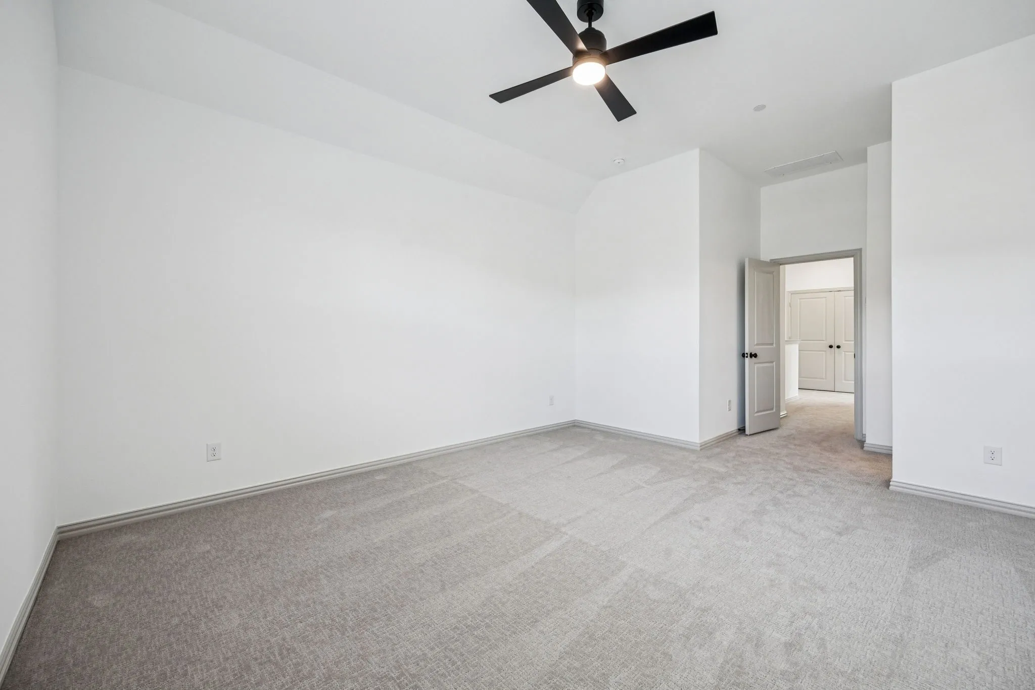 Spacious room featuring neutral carpeting, white walls, a black ceiling fan, and a light-colored interior door with dark hardware