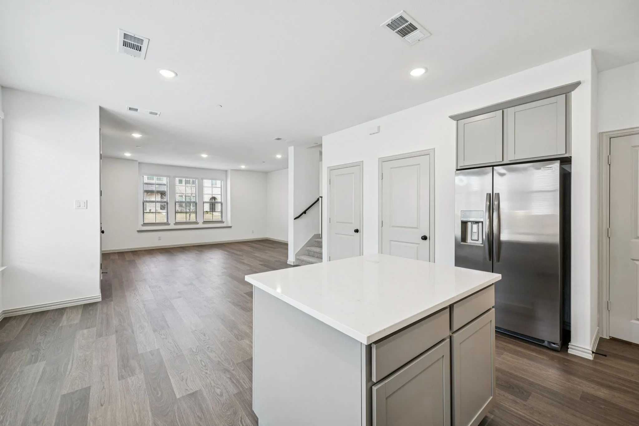 Open-concept living space with wood-finish flooring, a kitchen island with a light-colored countertop, recessed lighting, and a stainless steel refrigerator