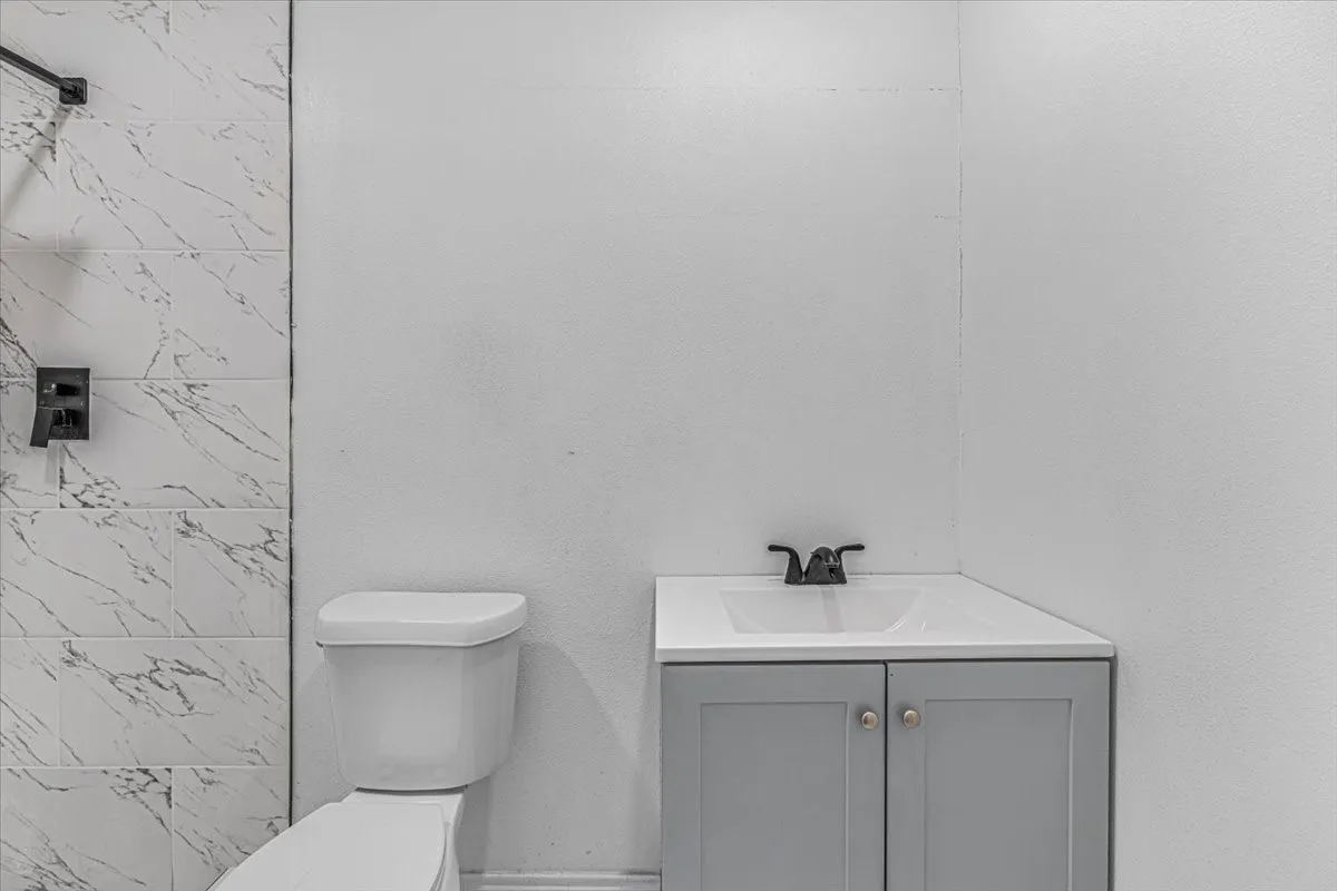 White ceramic toilet, corner vanity with gray cabinetry, white countertop, and black faucet, alongside a shower with white marble-look tile and black fixtures