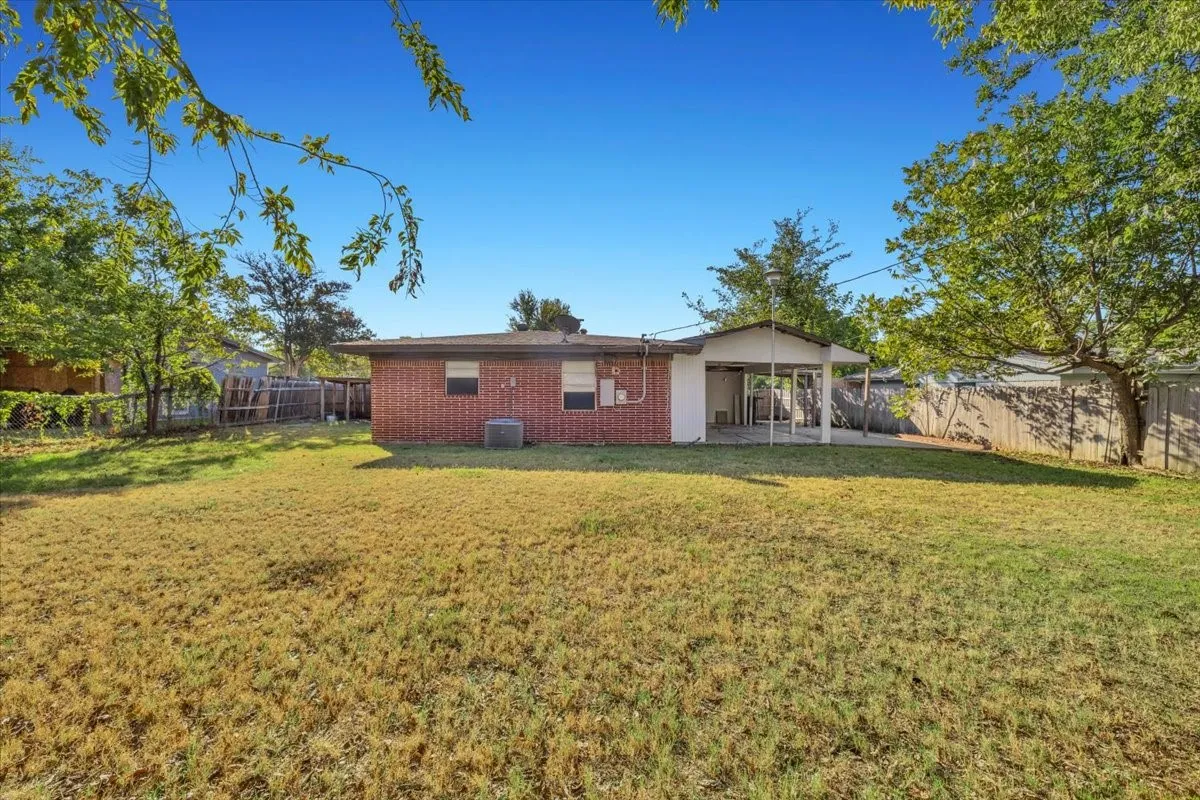 Expansive backyard with a covered patio, brick exterior, fenced perimeter, and mature trees
