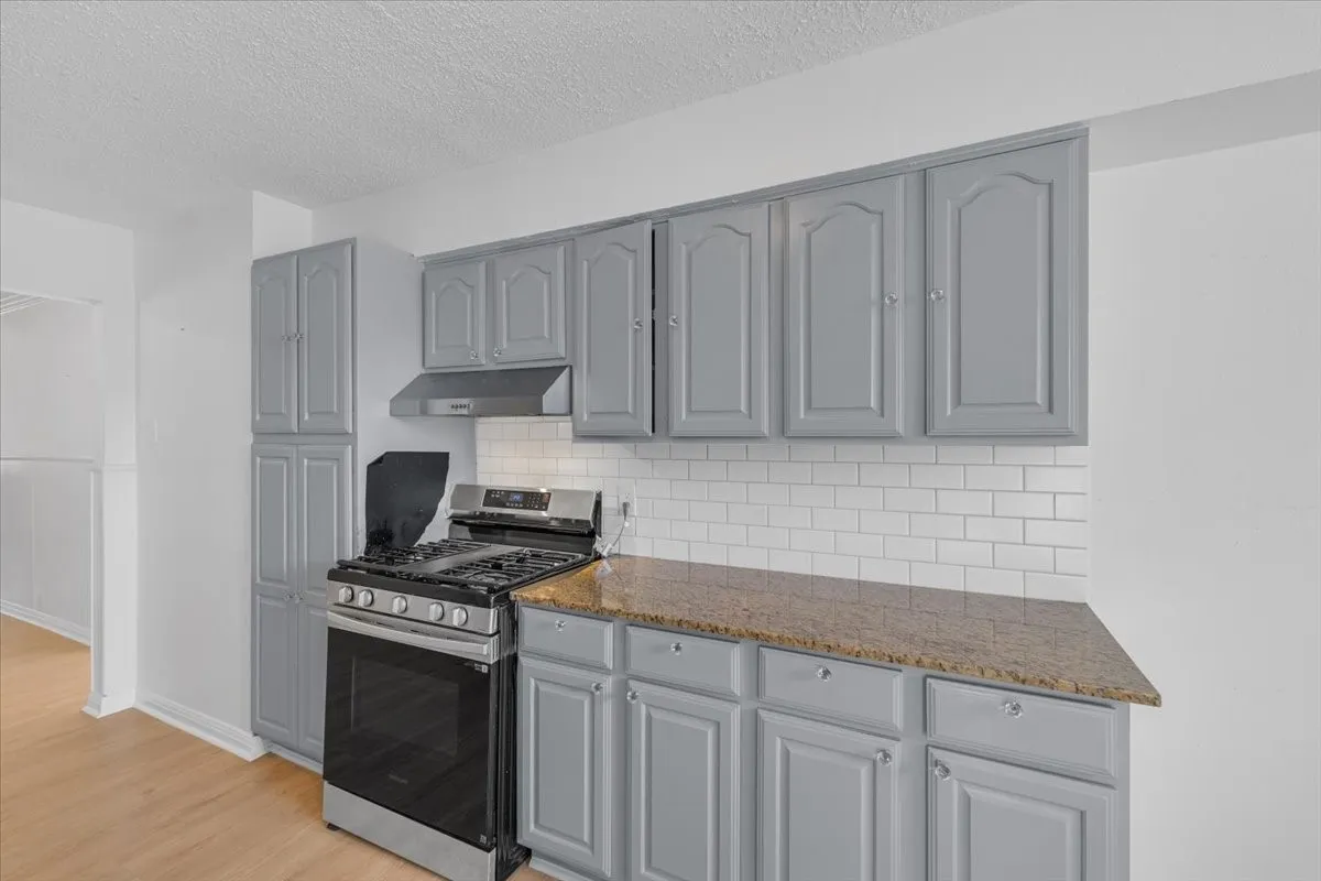 Stainless steel range with coordinating hood, white subway tile backsplash, light wood-finish flooring, grey cabinetry, and stone-finish countertops