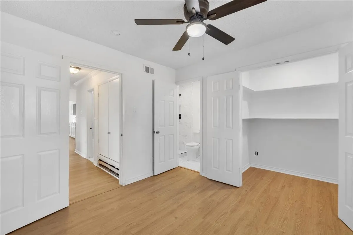 Wood-finish flooring, ceiling fan with integrated lighting, built-in shelving, white paneled doors, and a partial bathroom with a toilet and tiled walls