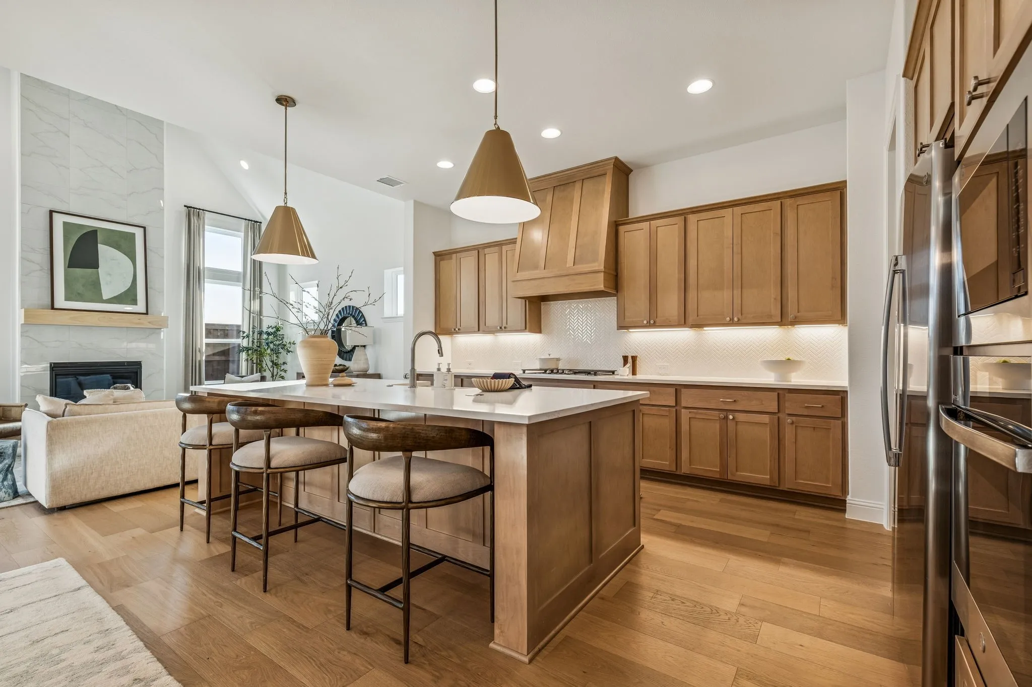 Open-concept living space featuring wood-finish flooring, a kitchen island with seating, pendant lighting, stainless steel appliances, and a fireplace with a stone surround