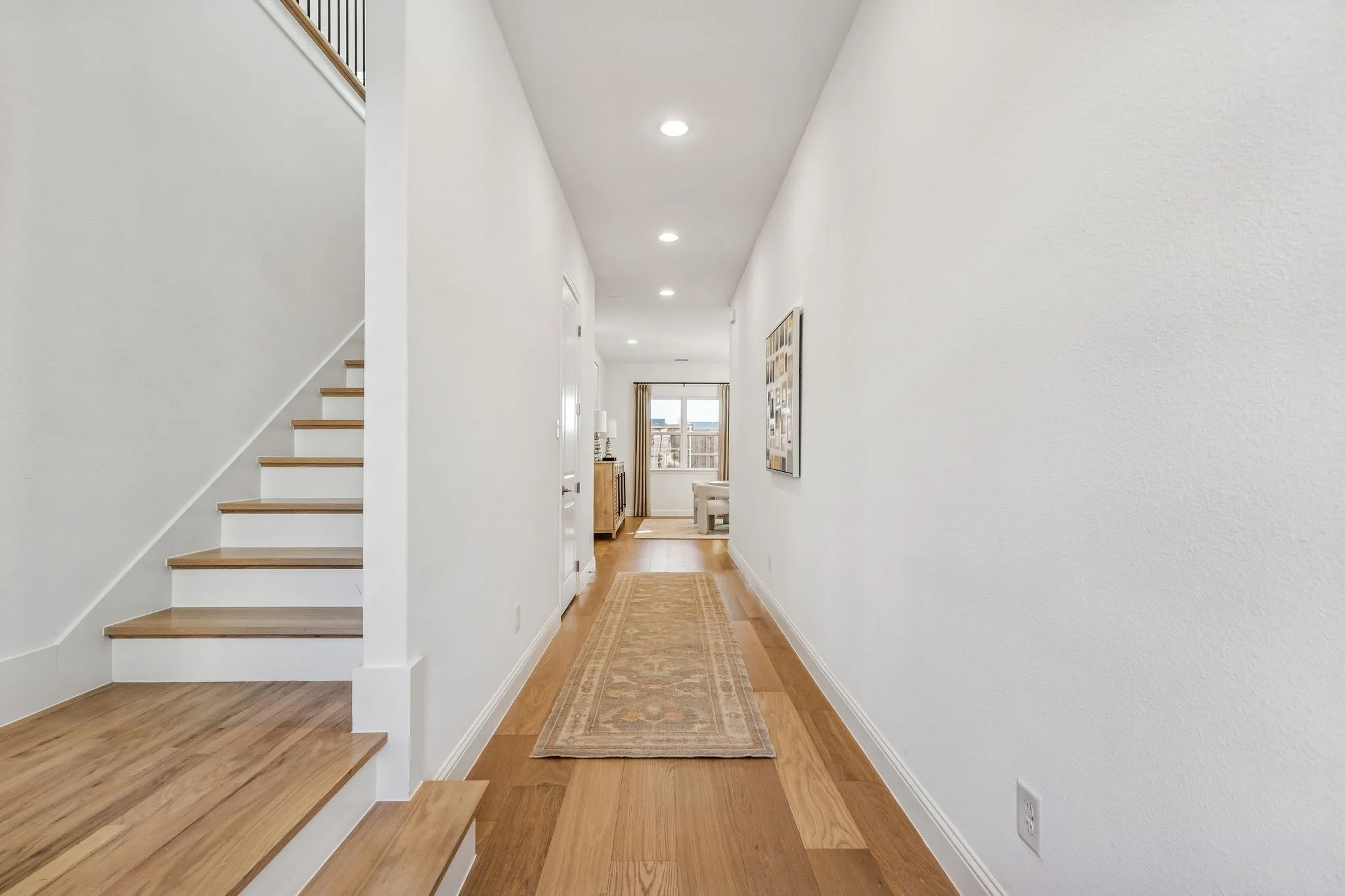 Wood-finish flooring throughout, white painted walls, recessed lighting, and a staircase with wood-finish treads and a dark metal railing