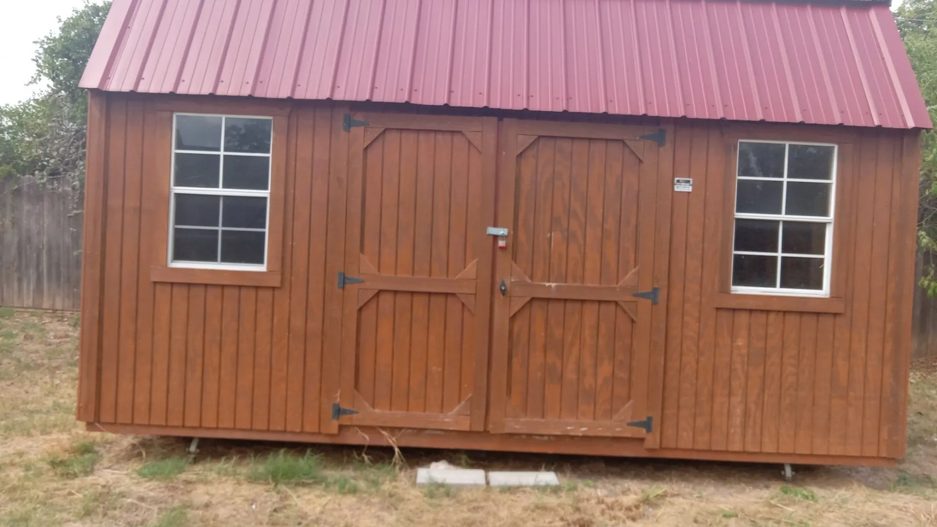 Wood-paneled storage shed featuring a red metal roof, double barn doors with black hardware, and two white-framed​​‌​​​​‌​​‌‌​‌‌​​​‌‌​‌​‌​‌​​​‌​​ windows