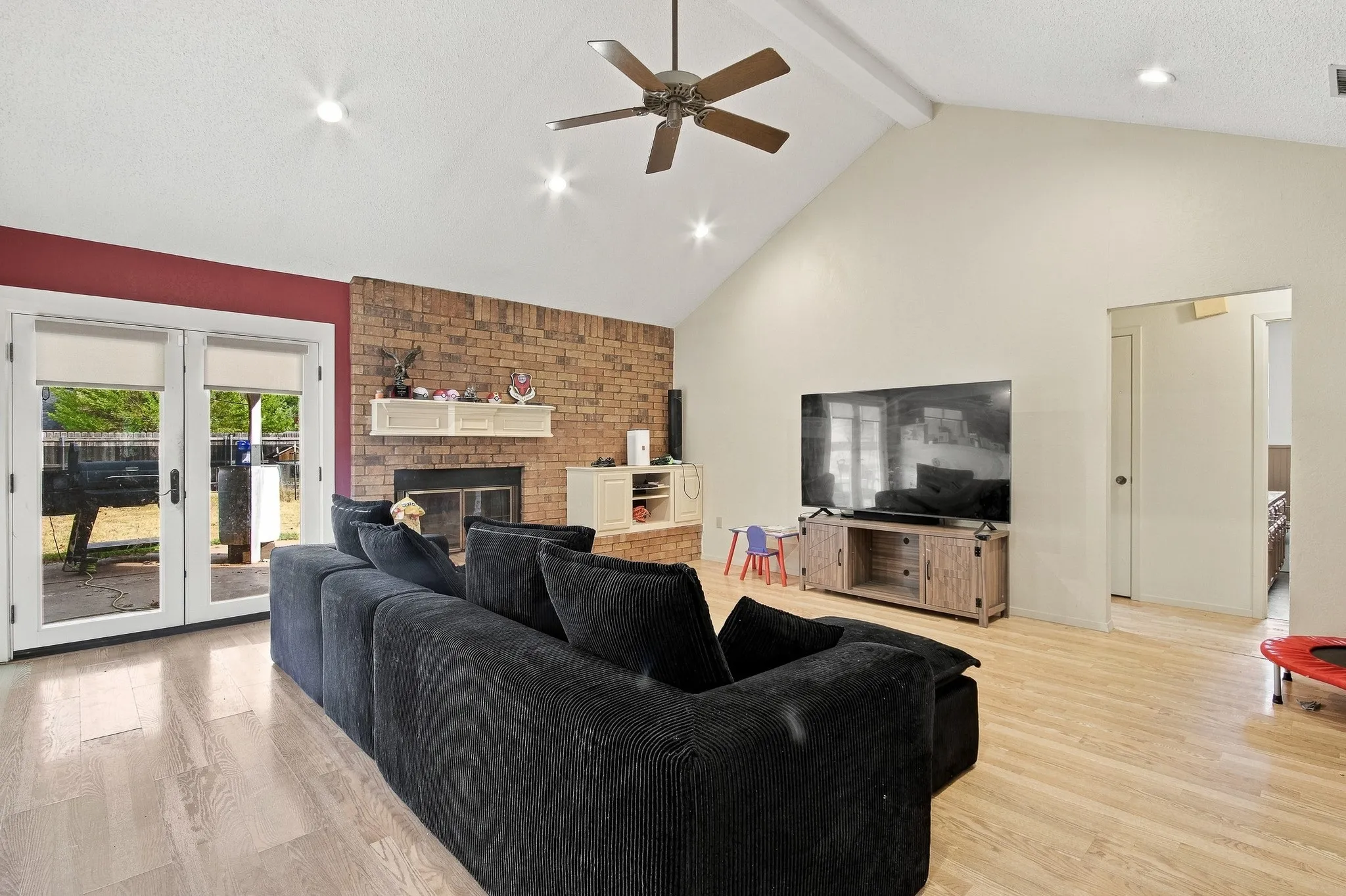 Vaulted ceiling, wood-finish flooring, brick-faced fireplace, white mantel, and ceiling fan with integrated lighting