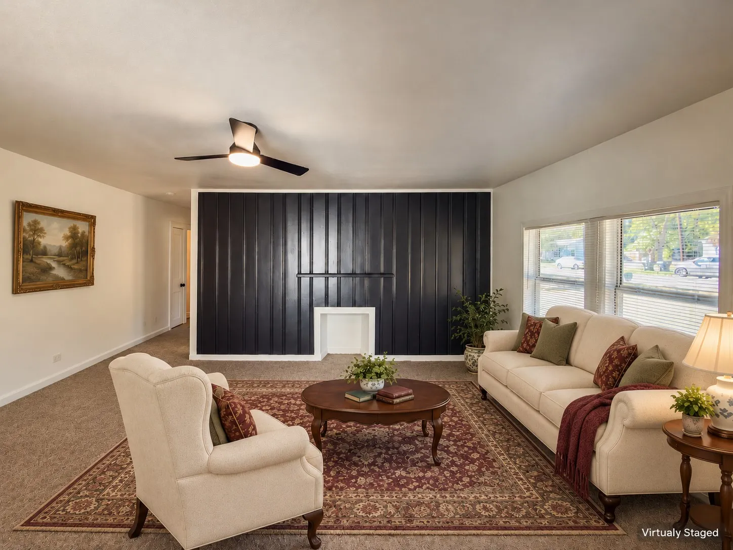 Spacious living room featuring a ceiling fan with integrated lighting, black vertical blinds, large windows with natural light exposure, cream-upholstered seating, wood-finish side tables, and patterned area rug over carpeted flooring