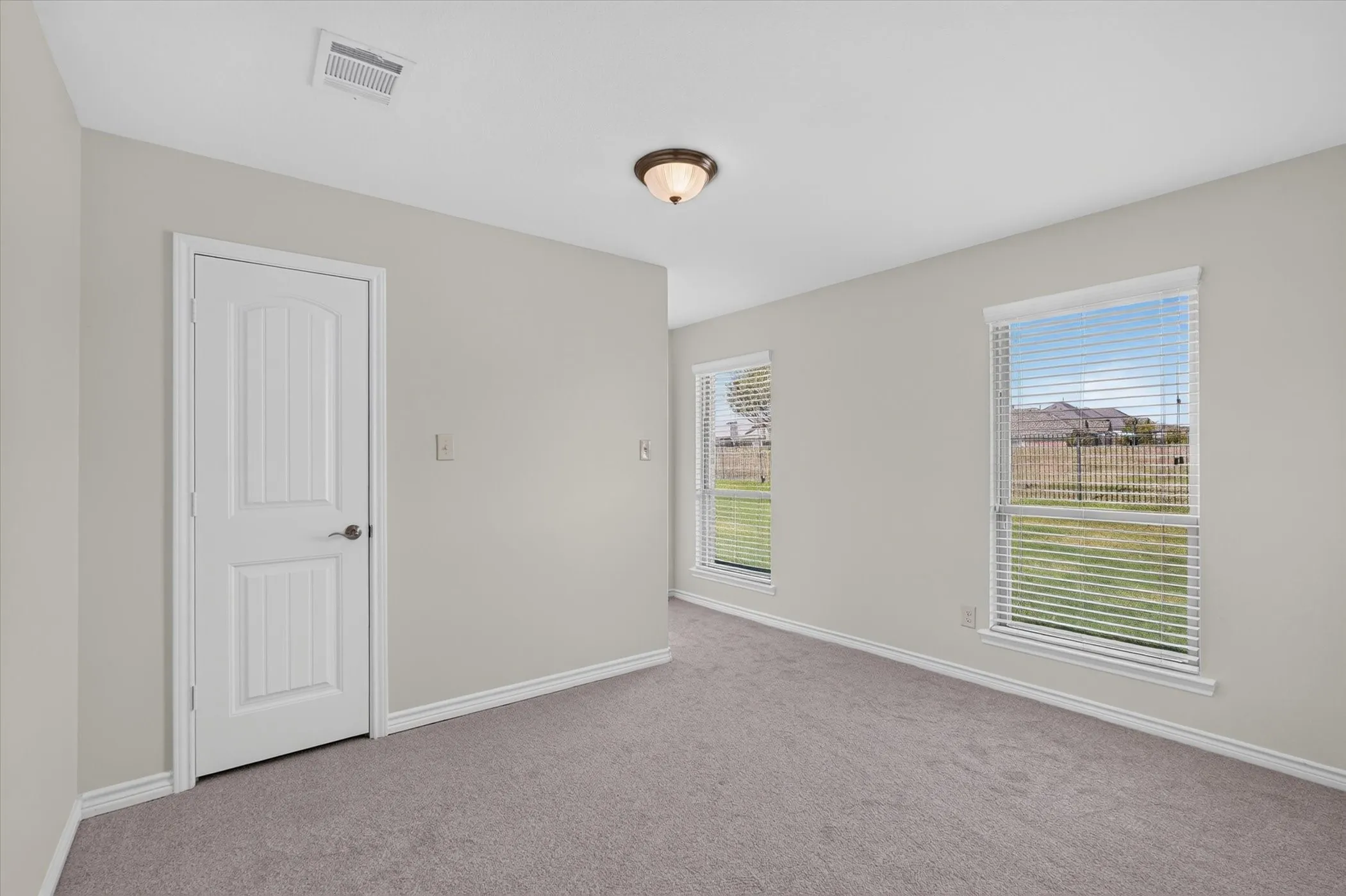 Soft gray carpeting with white baseboards, two windows with blinds, a white paneled door, and a flush mount ceiling fixture