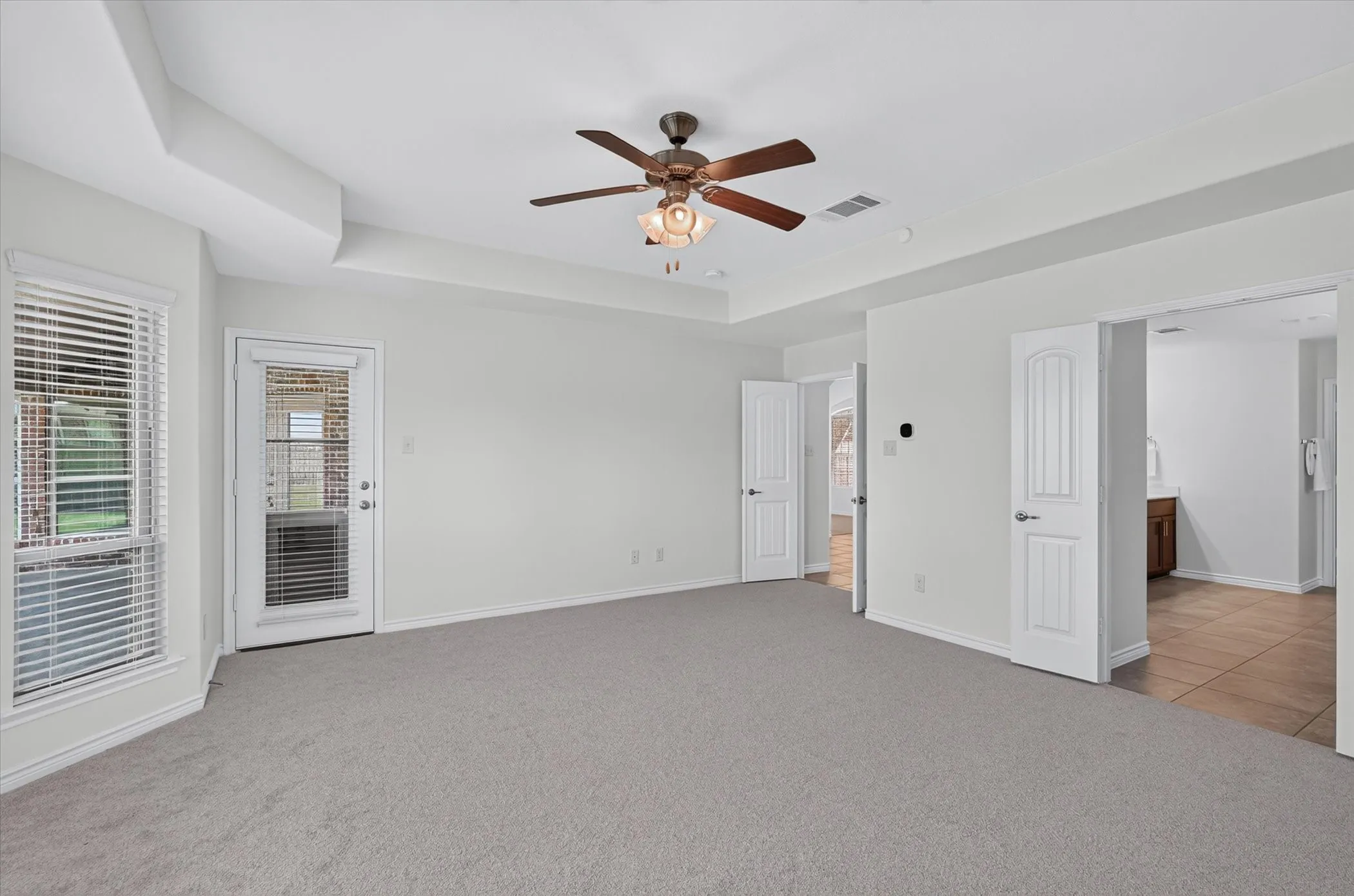 Tray ceiling with a ceiling fan, light gray carpeting, glass-paneled exterior door, and multiple interior doorways
