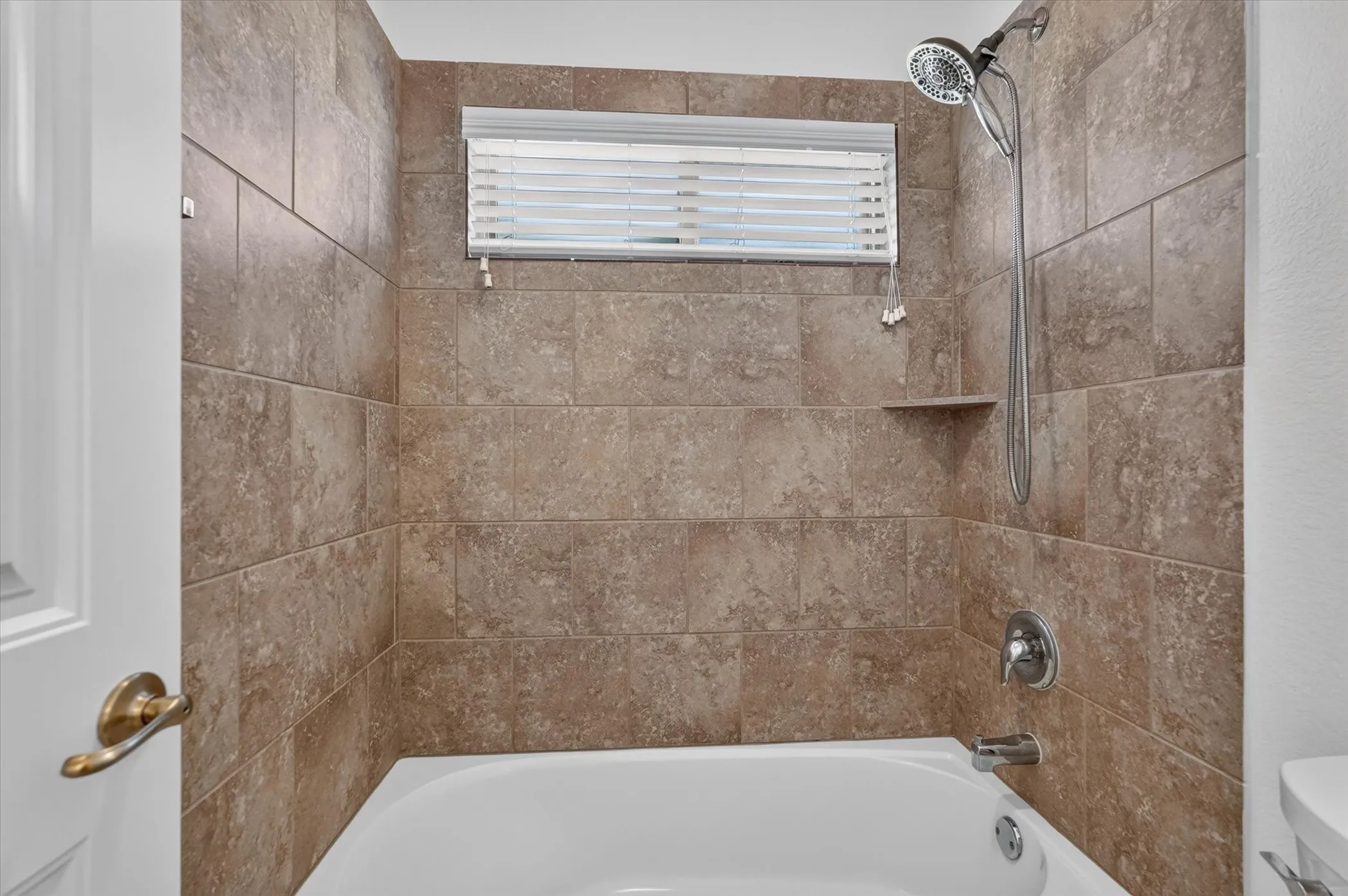 Bathtub with full-height tiled surround, integrated shower niche, and a window with blinds