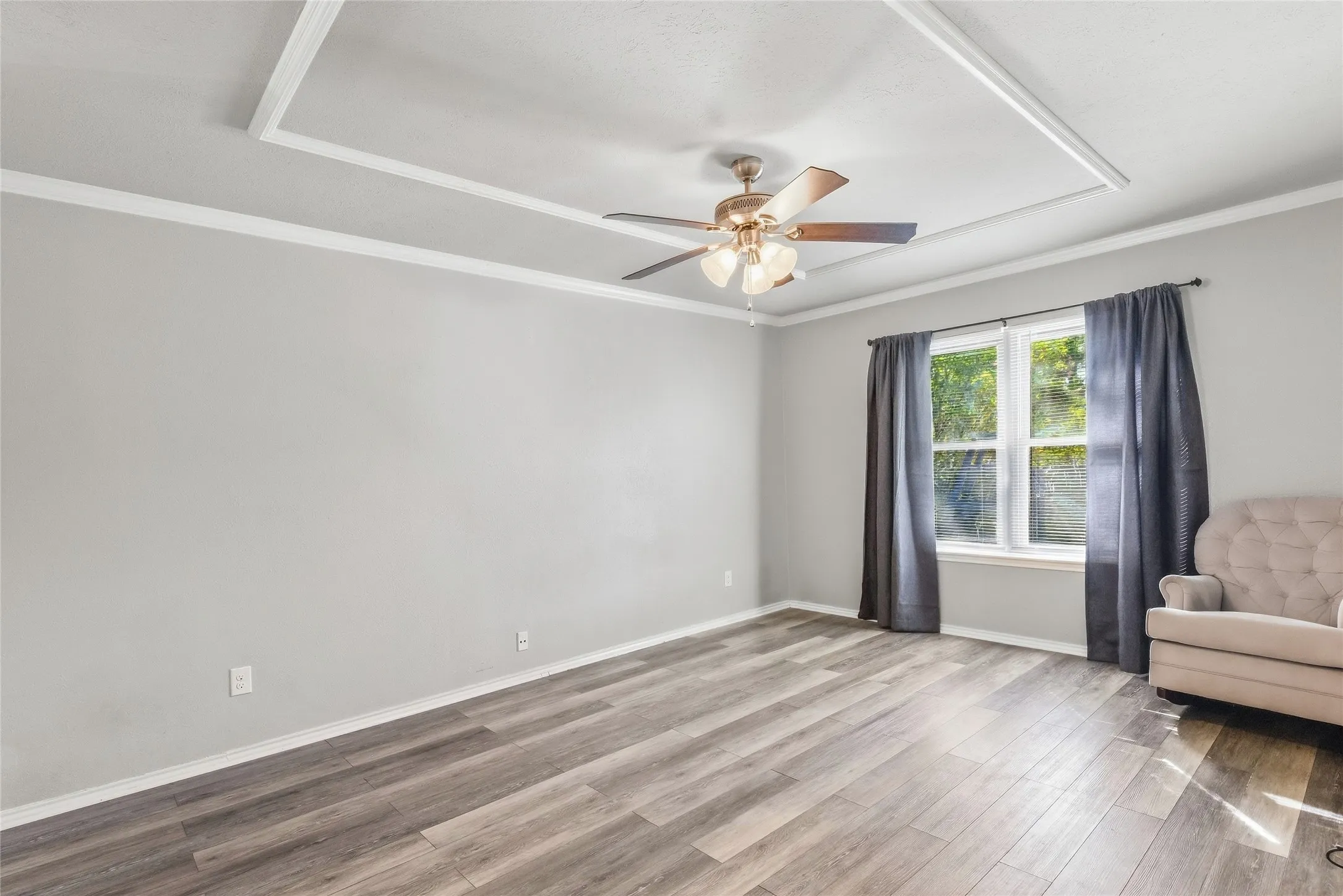 Wood-finish flooring, tray ceiling with crown molding, ceiling fan with light fixture, and window with trim