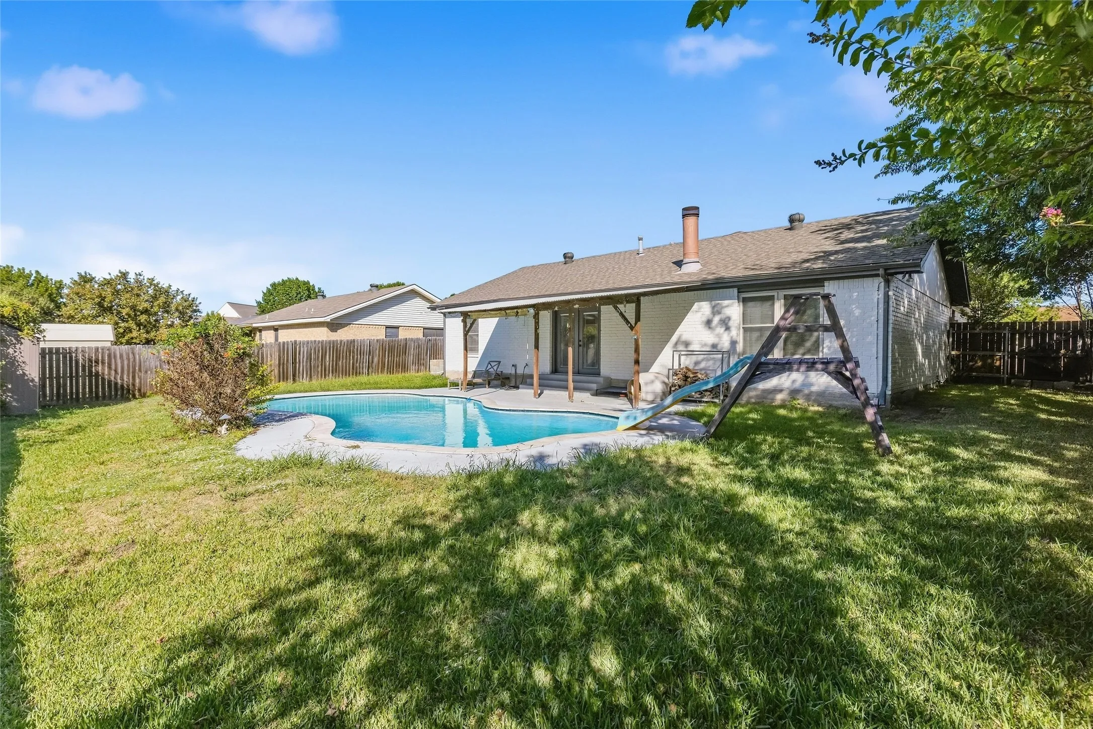 Expansive backyard featuring a built-in swimming pool with a surrounding paved deck, a covered patio with exposed wood beams, and a wood privacy fence