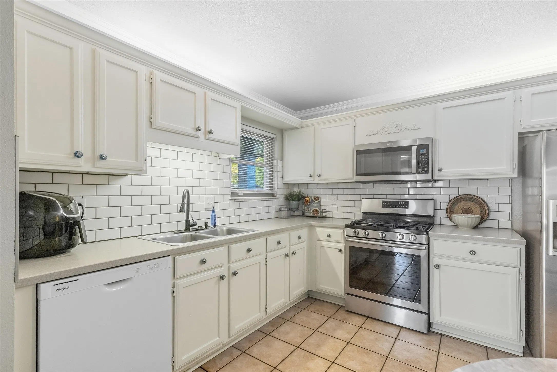White shaker-style cabinetry with crown molding, stainless steel appliances, subway tile backsplash, dual basin stainless steel sink, and light-toned countertops