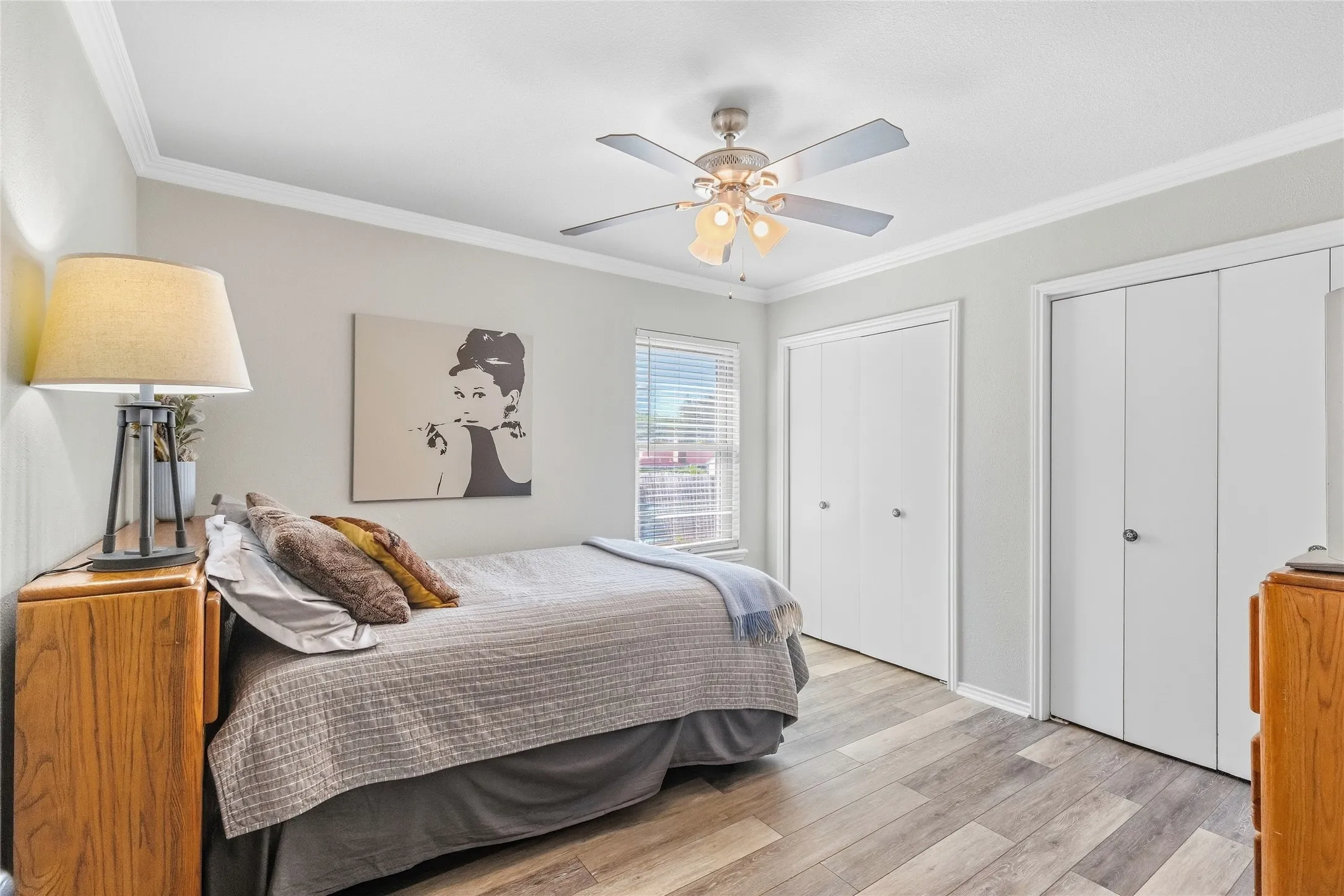 Light gray wall color, wood-finish flooring, two closet systems with white doors, ceiling fan with integrated lighting, and window with blinds