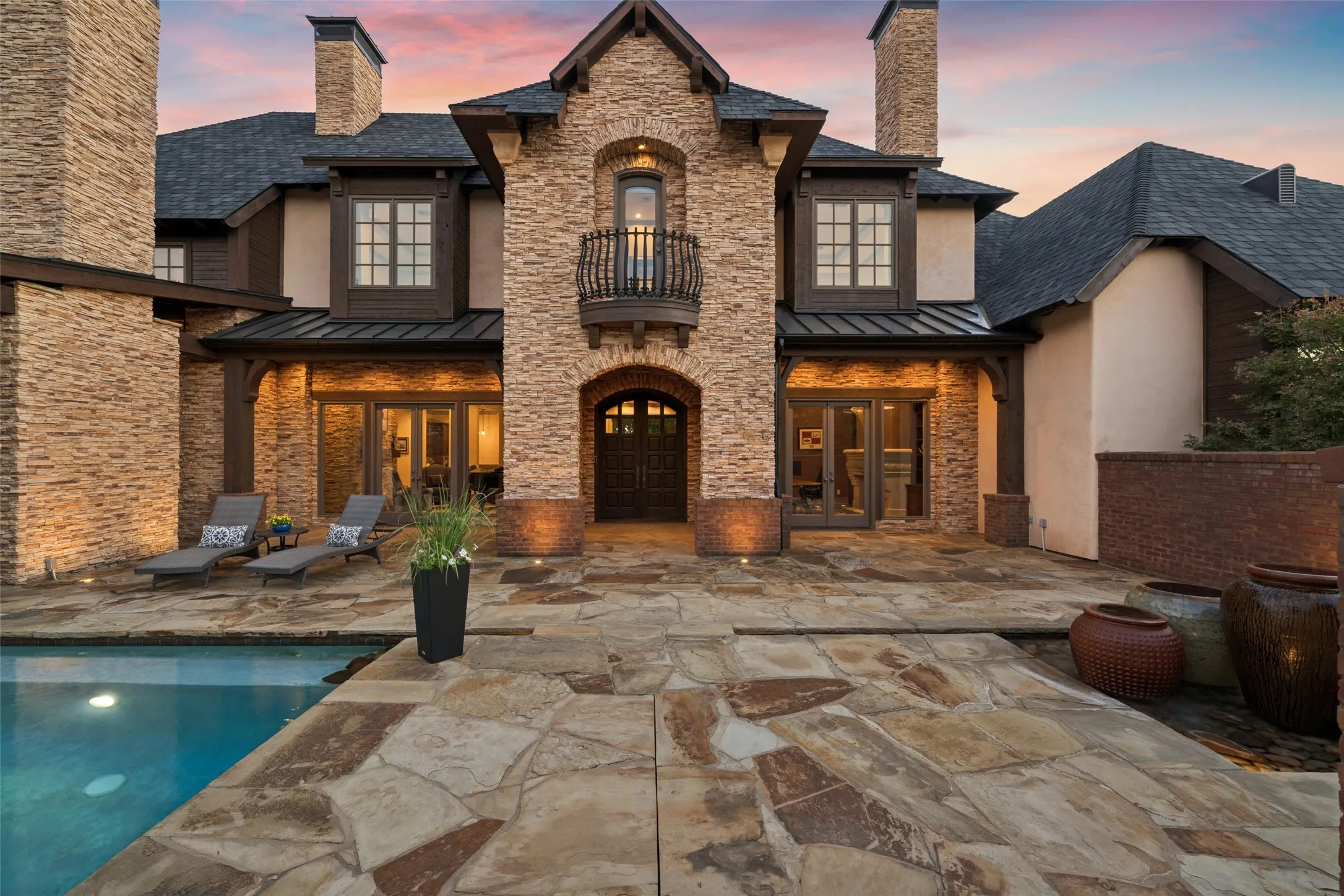 A commanding stacked-stone archway frames custom dark-wood double doors beneath a charming second-story Juliet balcony, creating an inviting, resort-style arrival