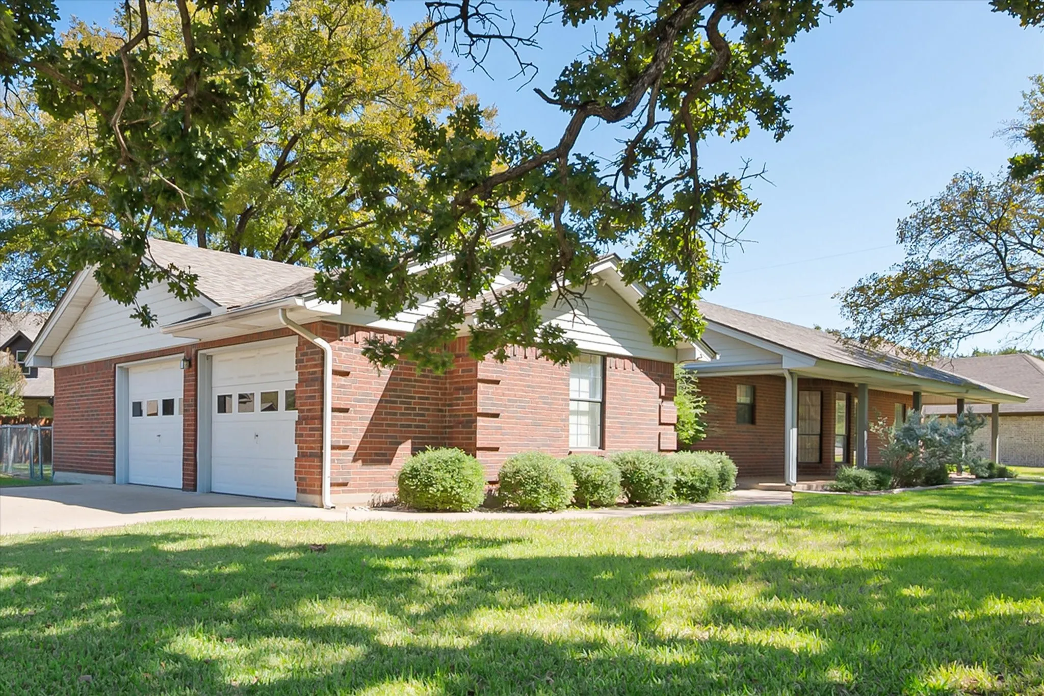 Brick exterior with white siding accents, two-car garage with panel doors, covered front porch, and mature trees providing shade