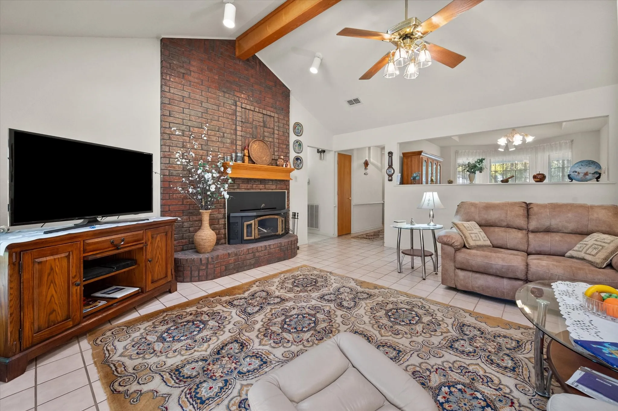 Vaulted ceiling with exposed wood beams, brick fireplace, tile flooring, and a ceiling fan with integrated lighting