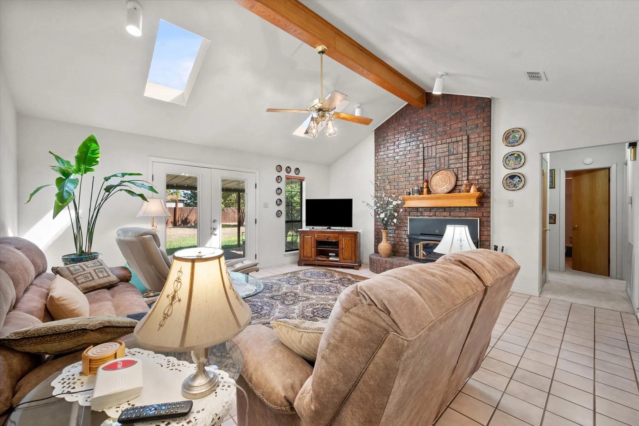 Vaulted ceiling with exposed wood beams, skylight, brick fireplace with wood mantel, ceiling fan with light fixture, and tile flooring
