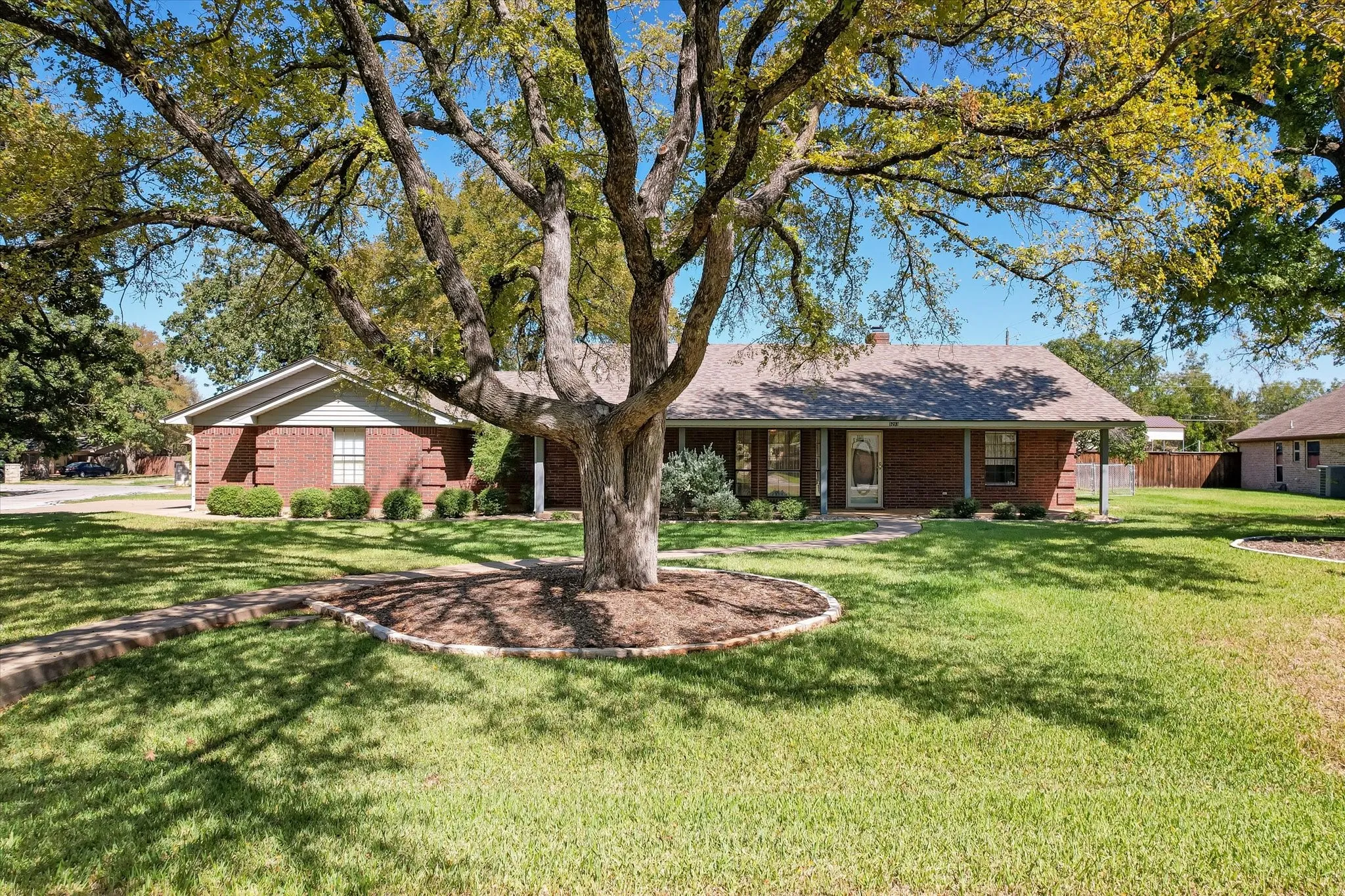 Brick exterior with a covered front porch, large mature tree, established landscaping, and a manicured lawn