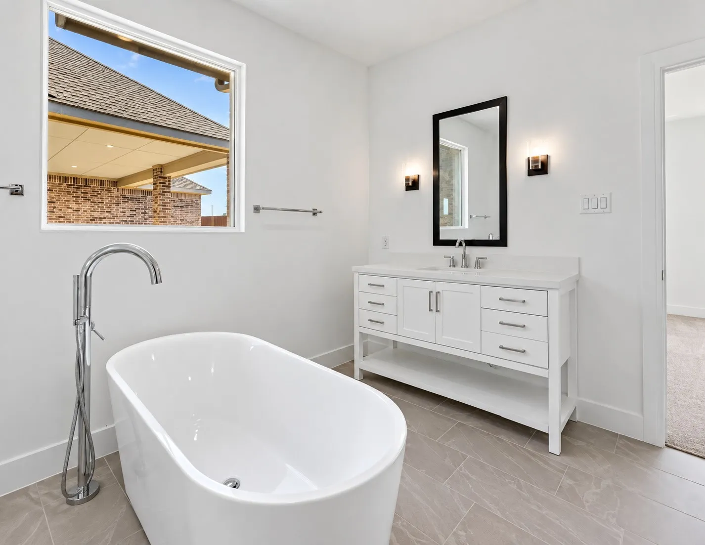 Freestanding soaking tub with a floor-mounted chrome faucet, white vanity with dual cabinet doors and multiple drawers, framed mirror, and wall-mounted sconces