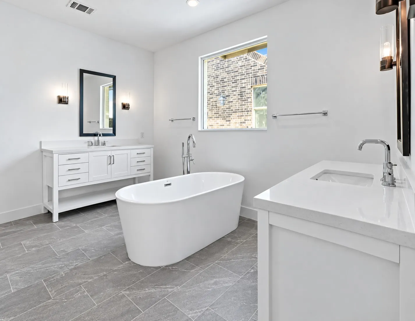 Freestanding soaking tub, dual white vanity cabinets, white countertops, chrome fixtures, and gray rectangular floor tiling
