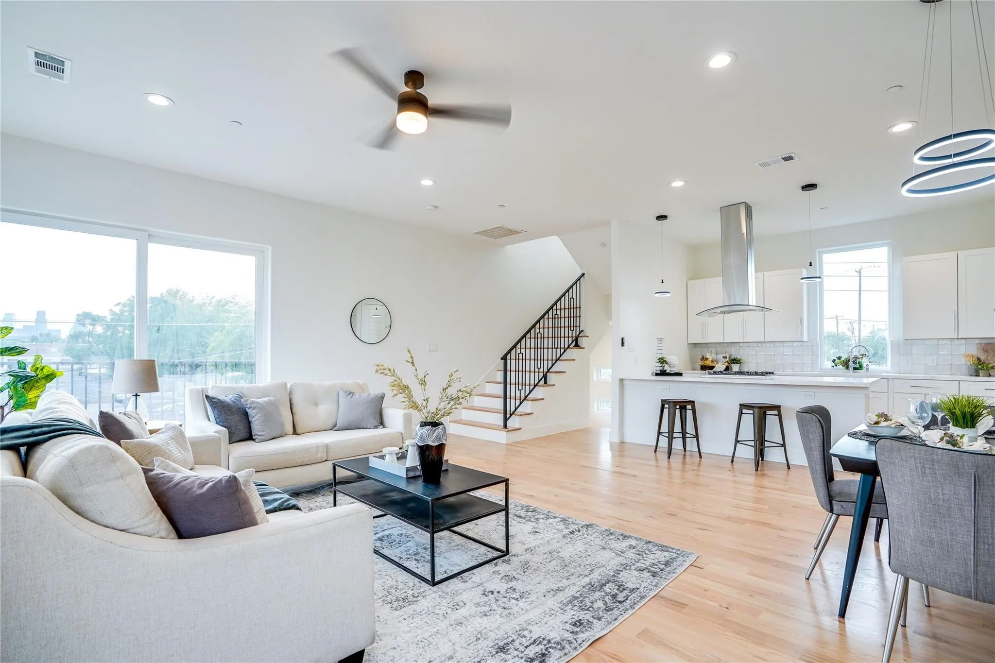 Open-concept living space featuring wood-finish flooring, a modern kitchen with a large island and stainless steel range hood, and a staircase with wrought iron balusters