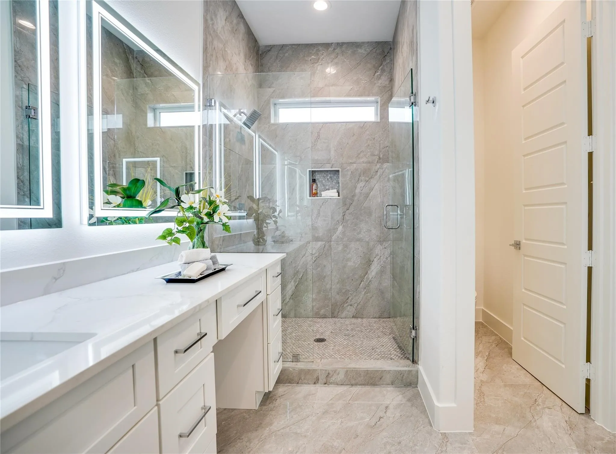 Modern bathroom featuring a double vanity with stone countertops and undermount sinks, frameless glass shower enclosure, large format tile flooring, and illuminated mirrors