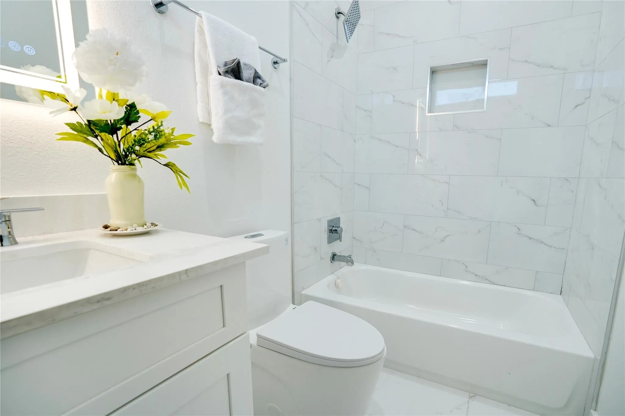 Bathroom featuring a white vanity with integrated sink and chrome faucet, illuminated mirror, white subway tile bathtub surround with recessed niche, and a modern toilet