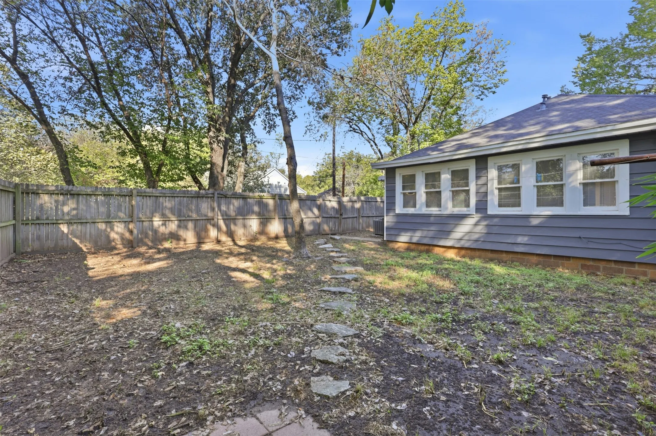 Fenced backyard featuring mature trees, a stepping stone path, and a gray siding exterior with white-framed windows