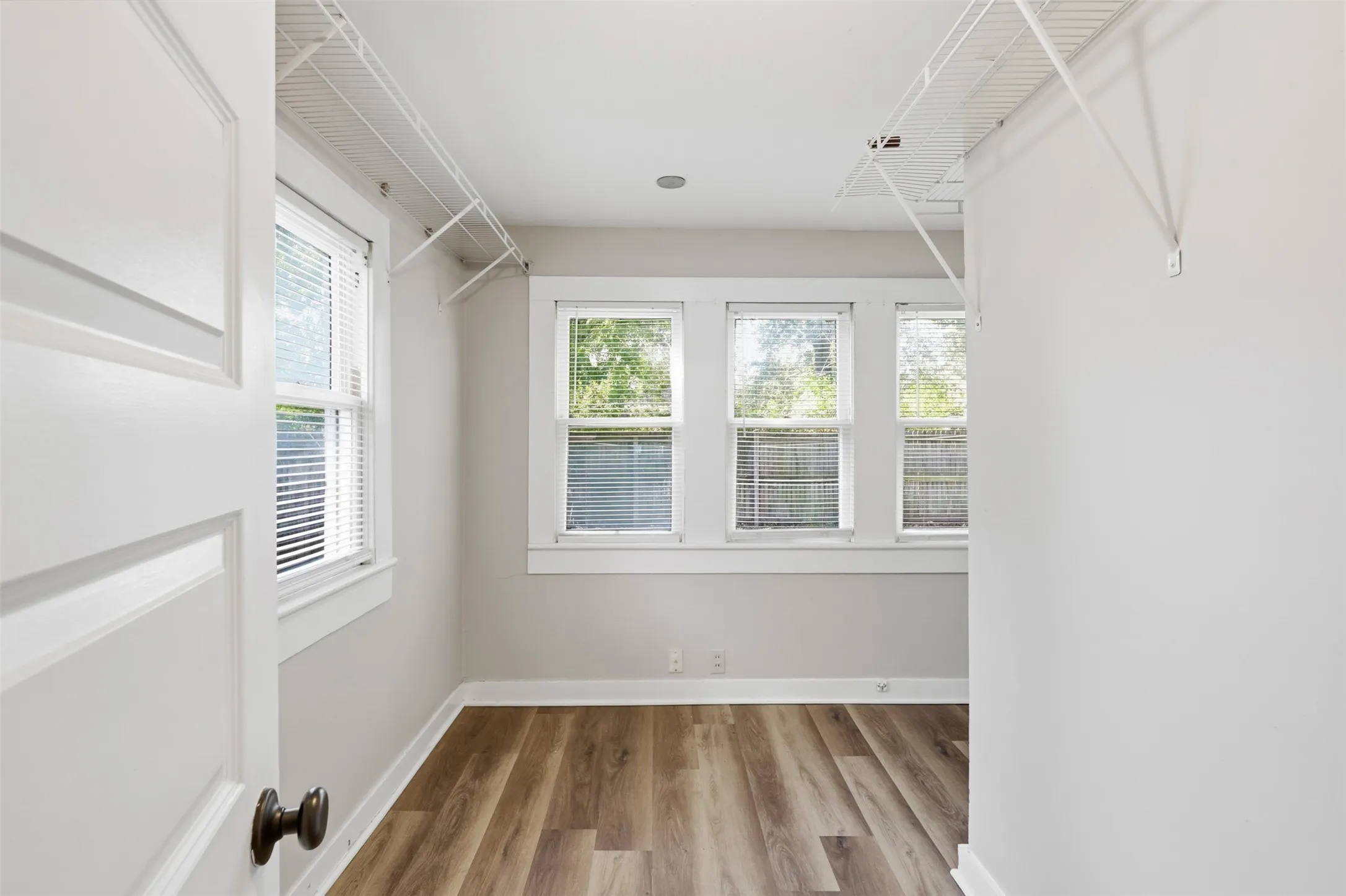 Light gray painted walls with white trim, wood-finish flooring, three windows with white blinds, and white wire shelving