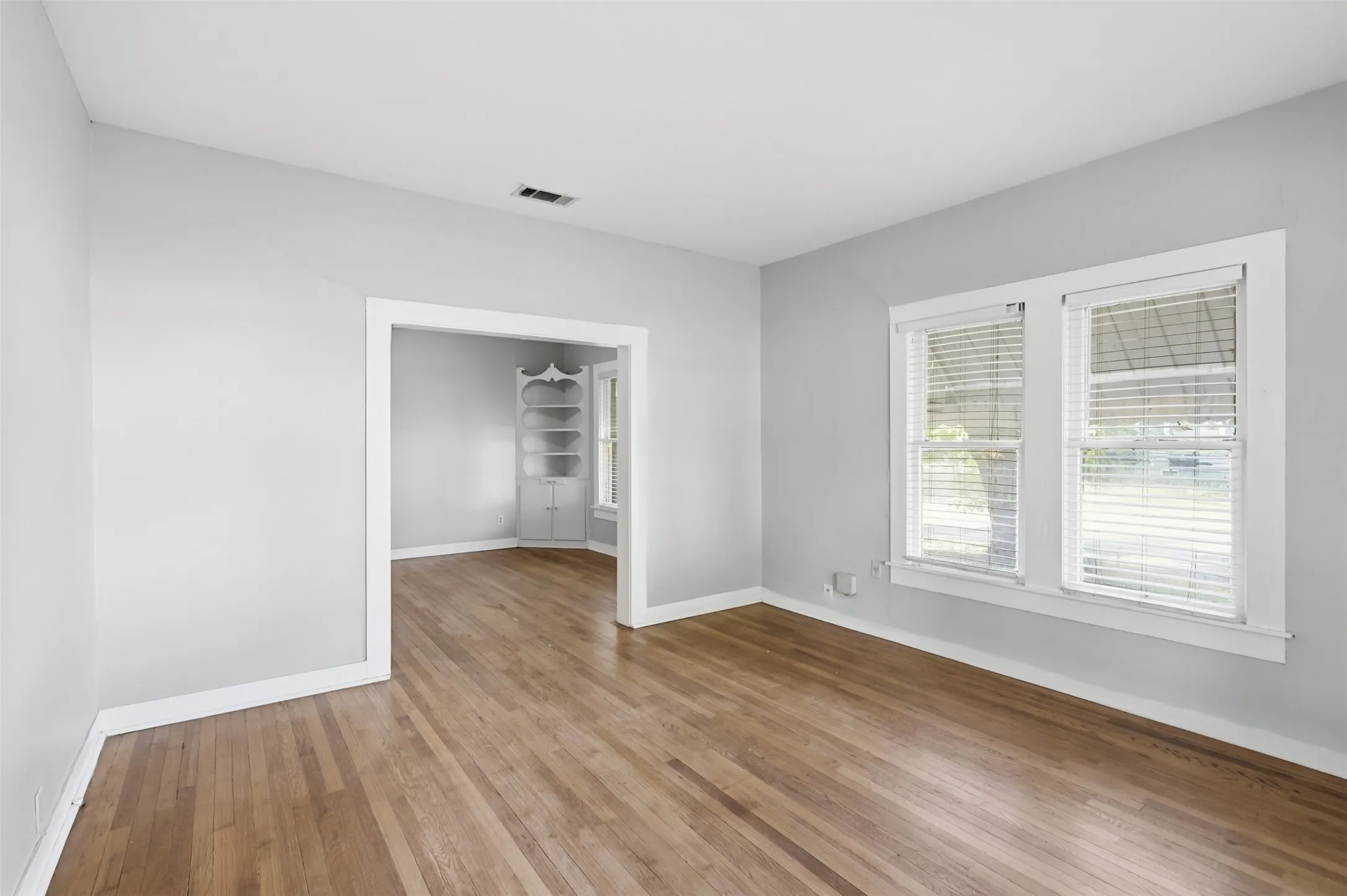 Hardwood flooring, painted walls, white trim, multiple windows with blinds, and a built-in corner cabinet