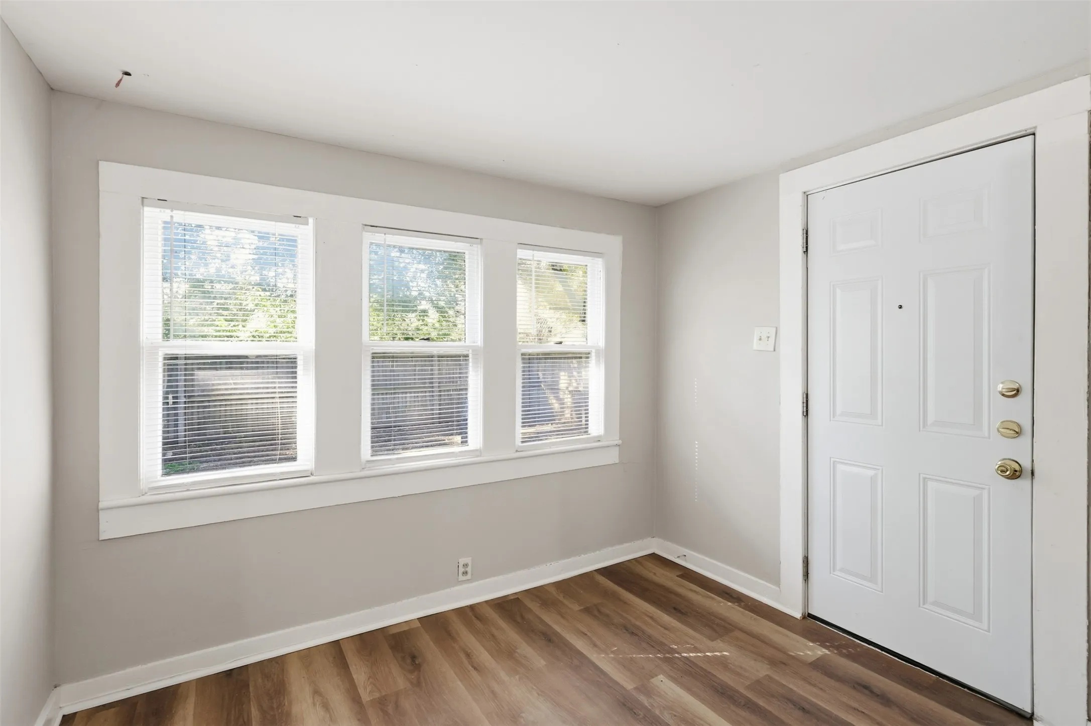 Wood-finish flooring, white paneled door with brass hardware, and three windows with white trim