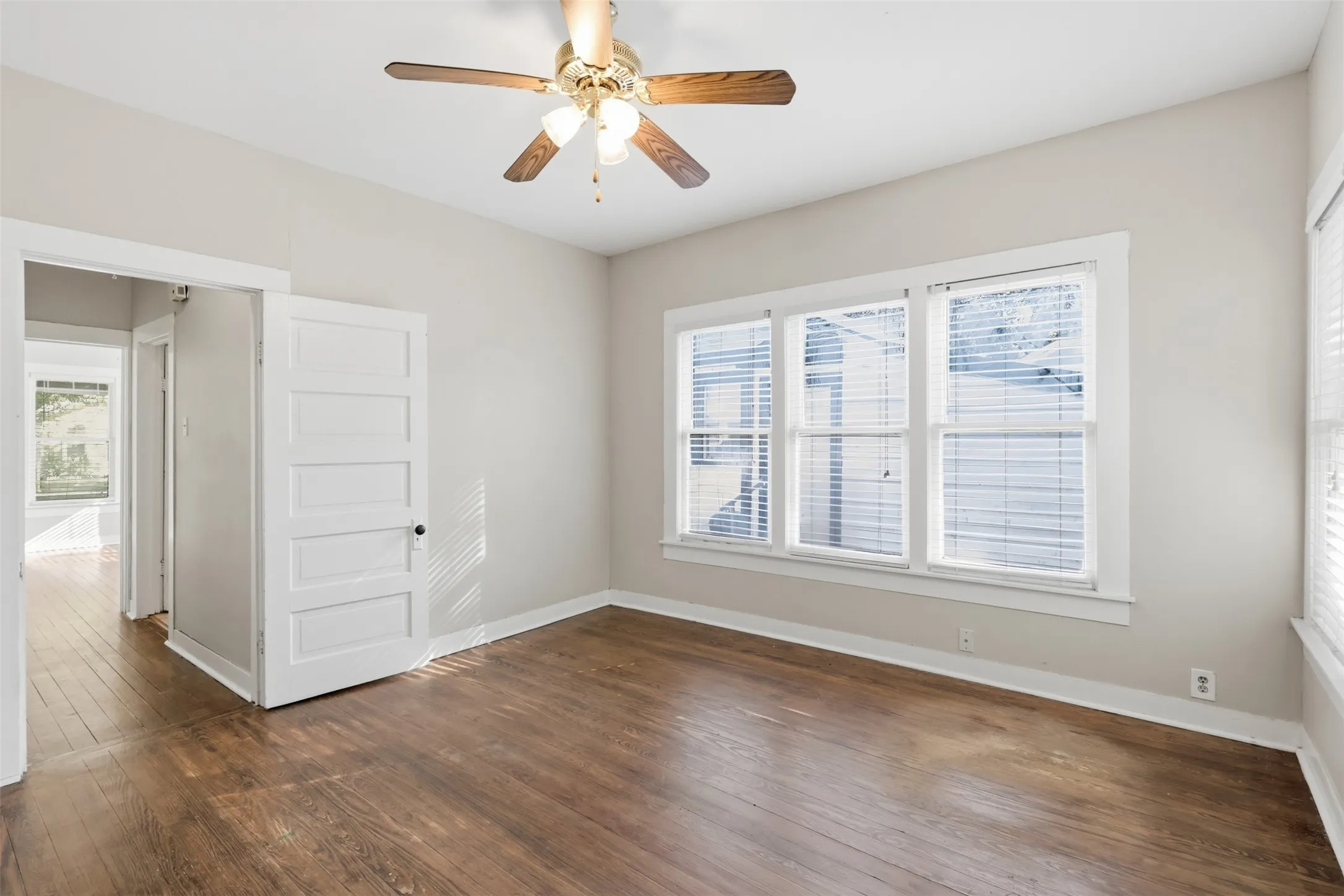Wood-finish flooring with crisp white baseboards, a classic five-panel door, multiple windows with blinds, and a ceiling fan with integrated lighting