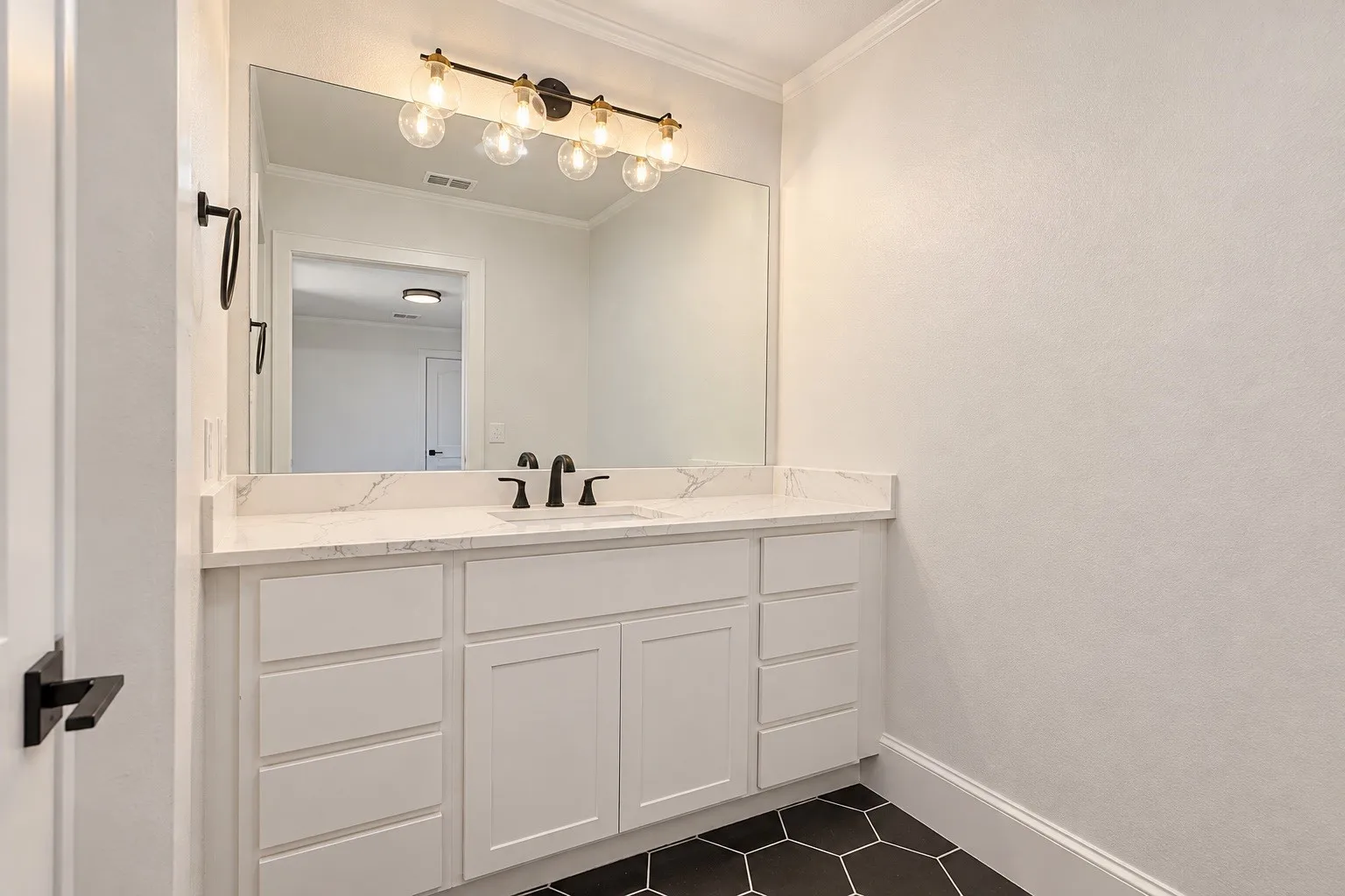 White vanity with multiple drawers, stone-finish countertop, black hexagonal floor tiles, large wall-mounted mirror, and a decorative light fixture