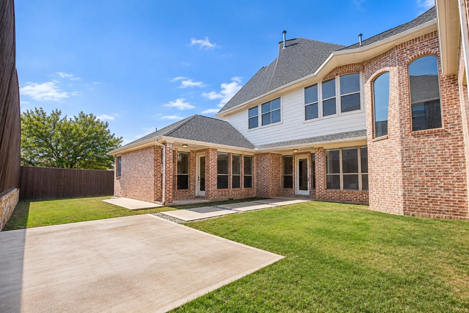 Expansive backyard featuring a manicured lawn, brick exterior, multiple concrete patios, and a privacy fence