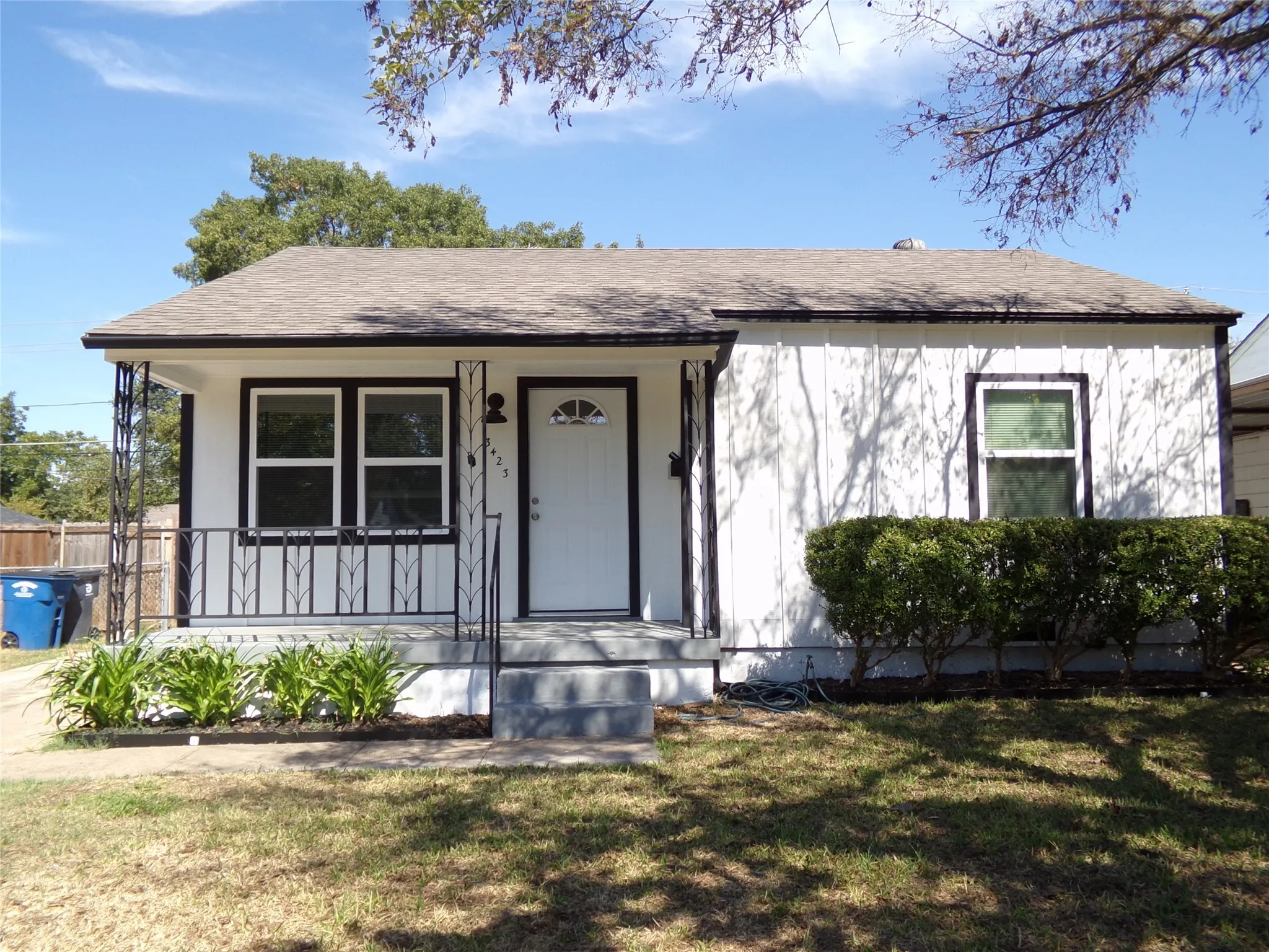 White exterior siding with dark trim, covered front entry with decorative metal railing, front door with arched lite window, and a planted shrub bed