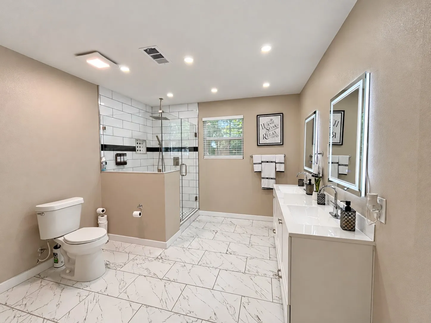 Dual vanity with integrated sinks, illuminated mirrors, glass-enclosed shower with subway tile, rainfall showerhead, and white-finish tile flooring
