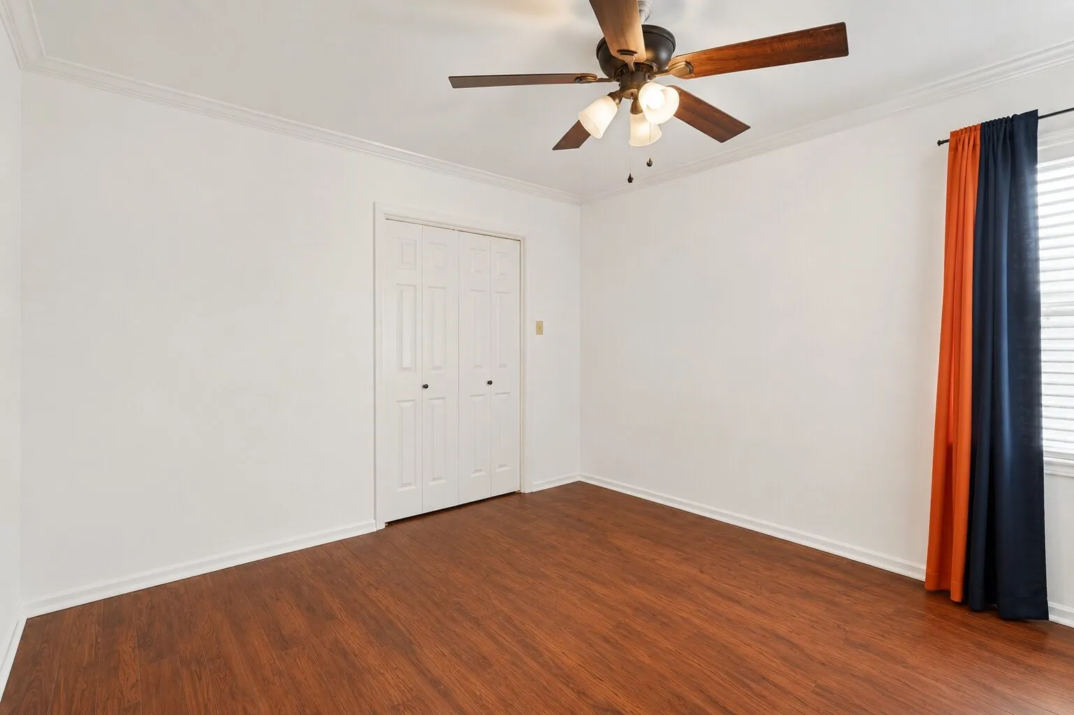 Wood-finish flooring, crown molding, ceiling fan with integrated lighting, bi-fold closet doors, and window with blinds