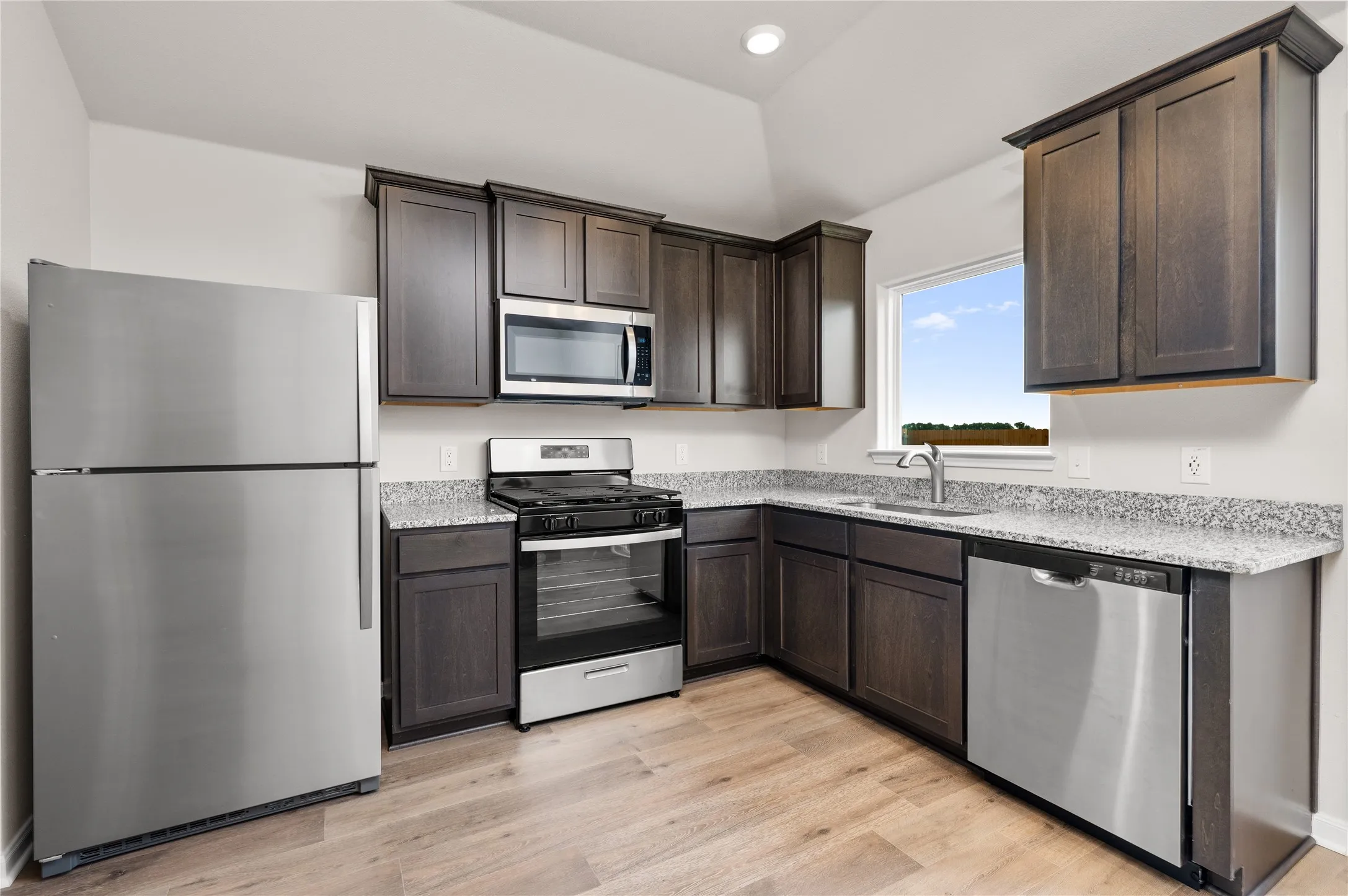 Kitchen with stainless steel appliances, light wood-style flooring, dark brown cabinets, light stone countertops, and recessed lighting