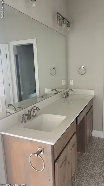 Dual undermount sinks with brushed nickel faucets, expansive vanity with light-toned wood-finish cabinetry, full-width mirror, and patterned floor tile