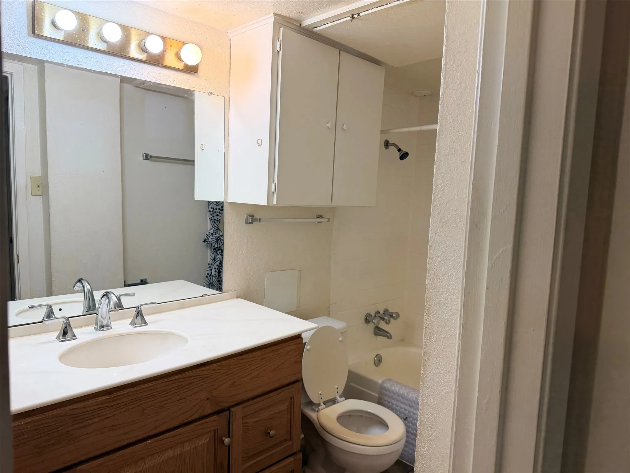 Bathroom featuring a wood-finish vanity with an integrated sink and chrome faucet, a large mirrored wall, overhead cabinetry, and a bathtub with an integrated shower head