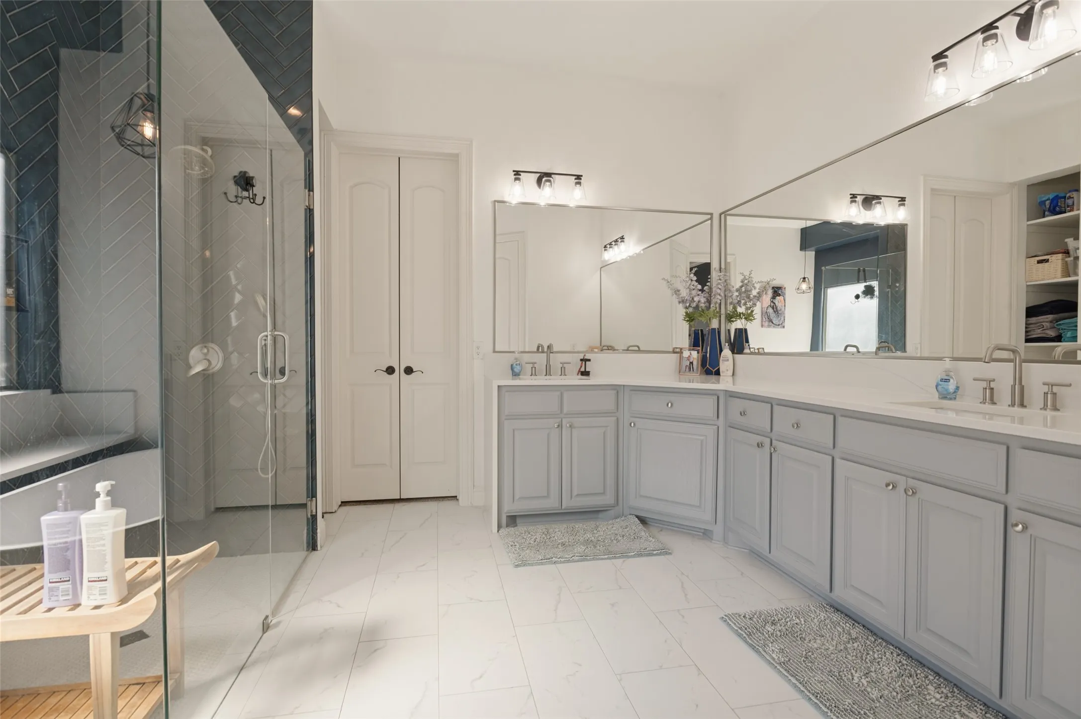 Spacious bathroom featuring an expansive dual vanity with light gray cabinetry and white countertops, a large frameless glass shower enclosure, and white tile flooring
