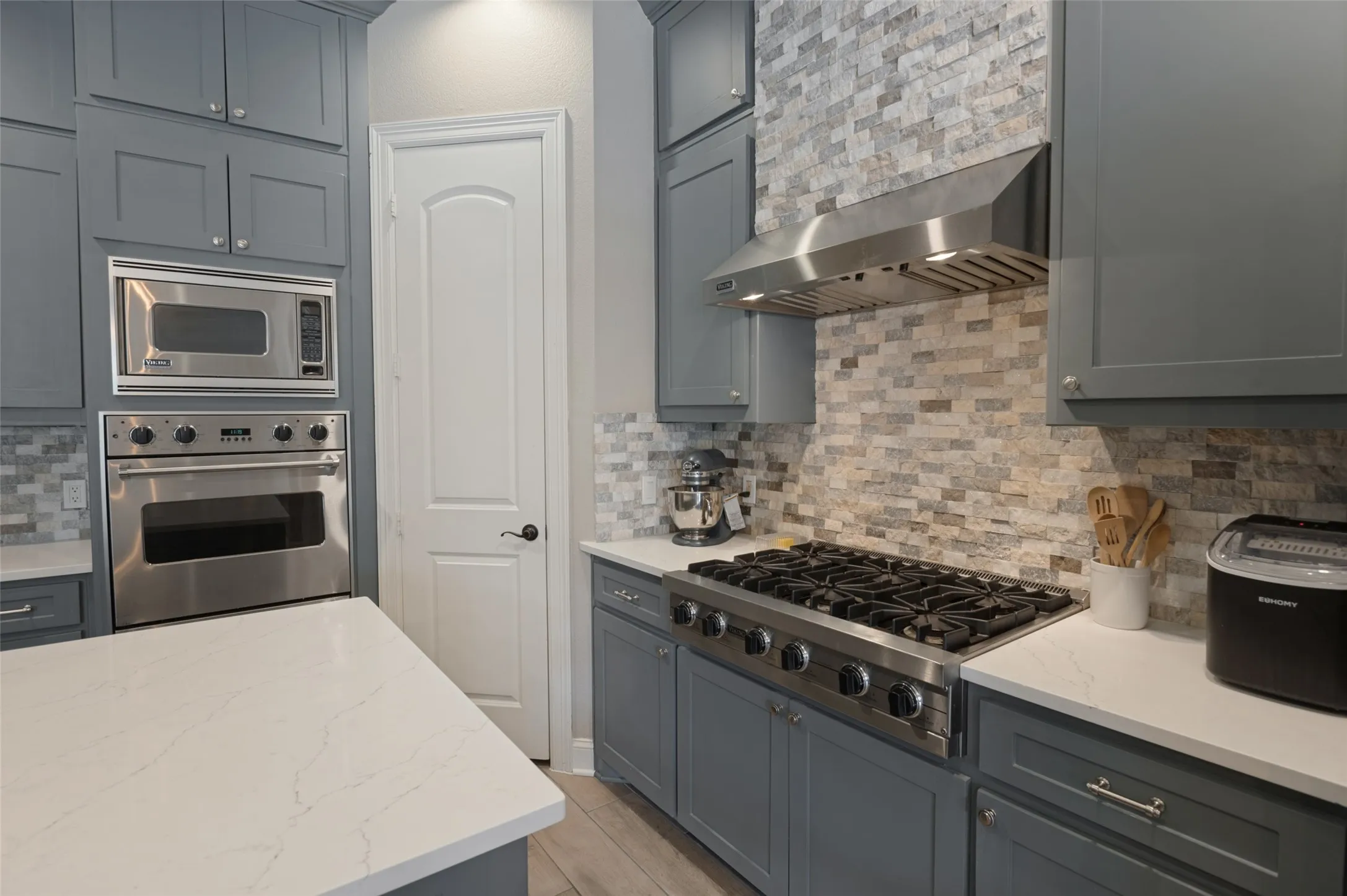 Gray cabinetry, built-in stainless steel oven and microwave, professional-grade gas range with stainless steel hood, quartz countertops, and a stone tile backsplash
