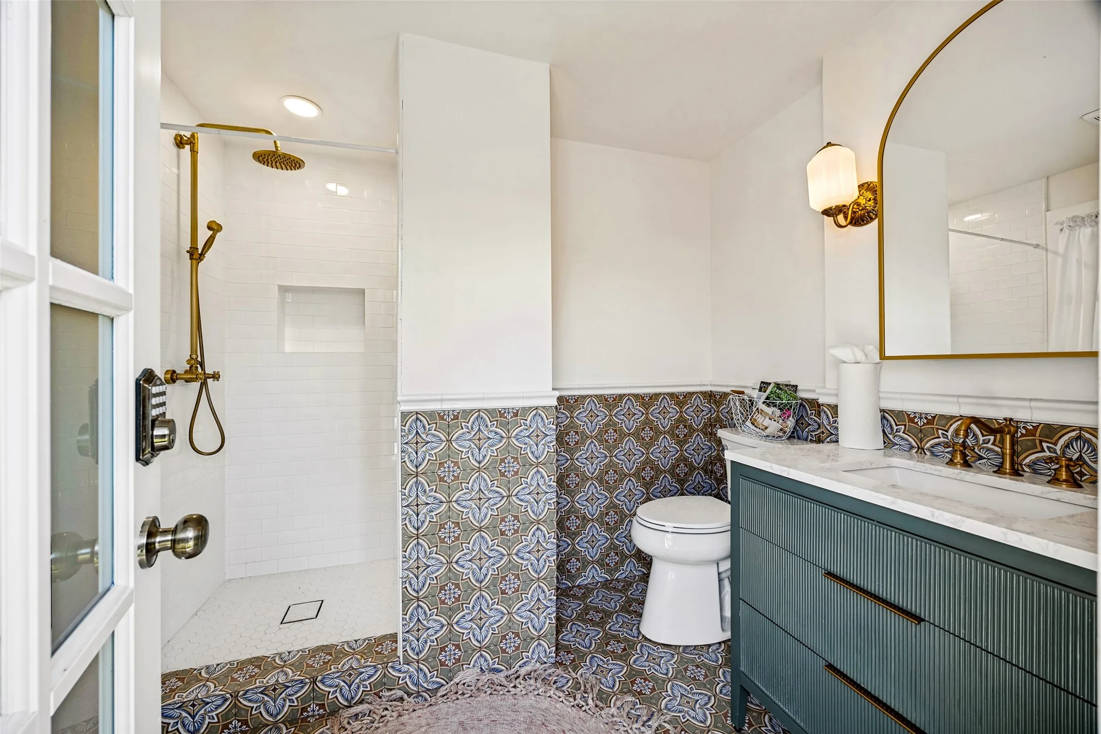 Walk-in shower with brass fixtures and recessed niche, patterned floor and wall tile, vanity with integrated sink and storage, arched mirror with sconce lighting