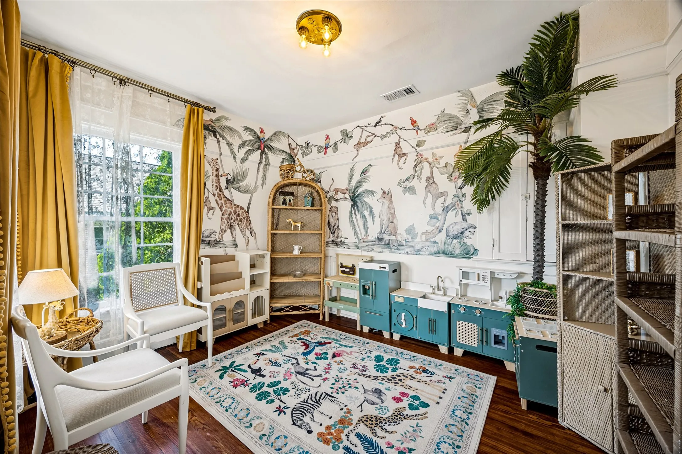 Bright corner study featuring hardwood flooring, custom built-in shelving with integrated storage, botanical wallcovering, large multi-pane windows with gold drapery hardware, and a brass ceiling fixture