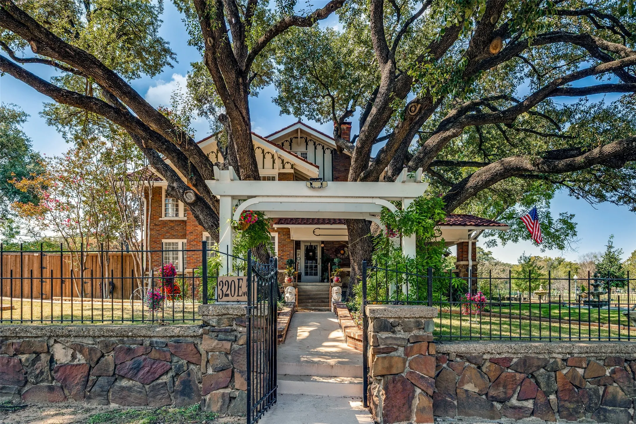 Brick exterior with white trim, mature trees, stone and wrought iron fencing, concrete walkway, and a white pergola with decorative details