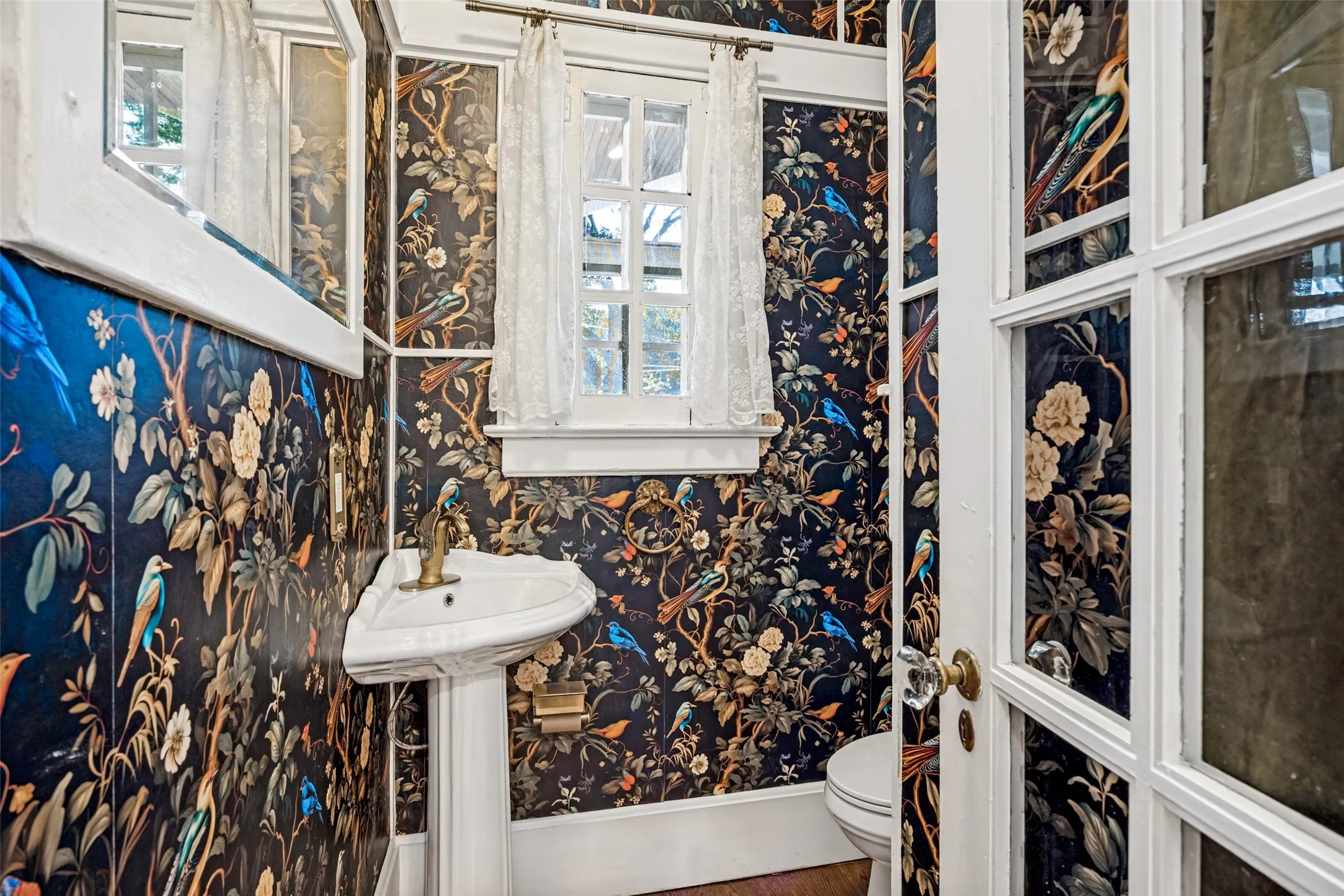Powder room featuring bold botanical wallcovering in navy, gold, and blue tones, pedestal sink with chrome hardware, white painted window casement with multiple panes, and wood-finish flooring