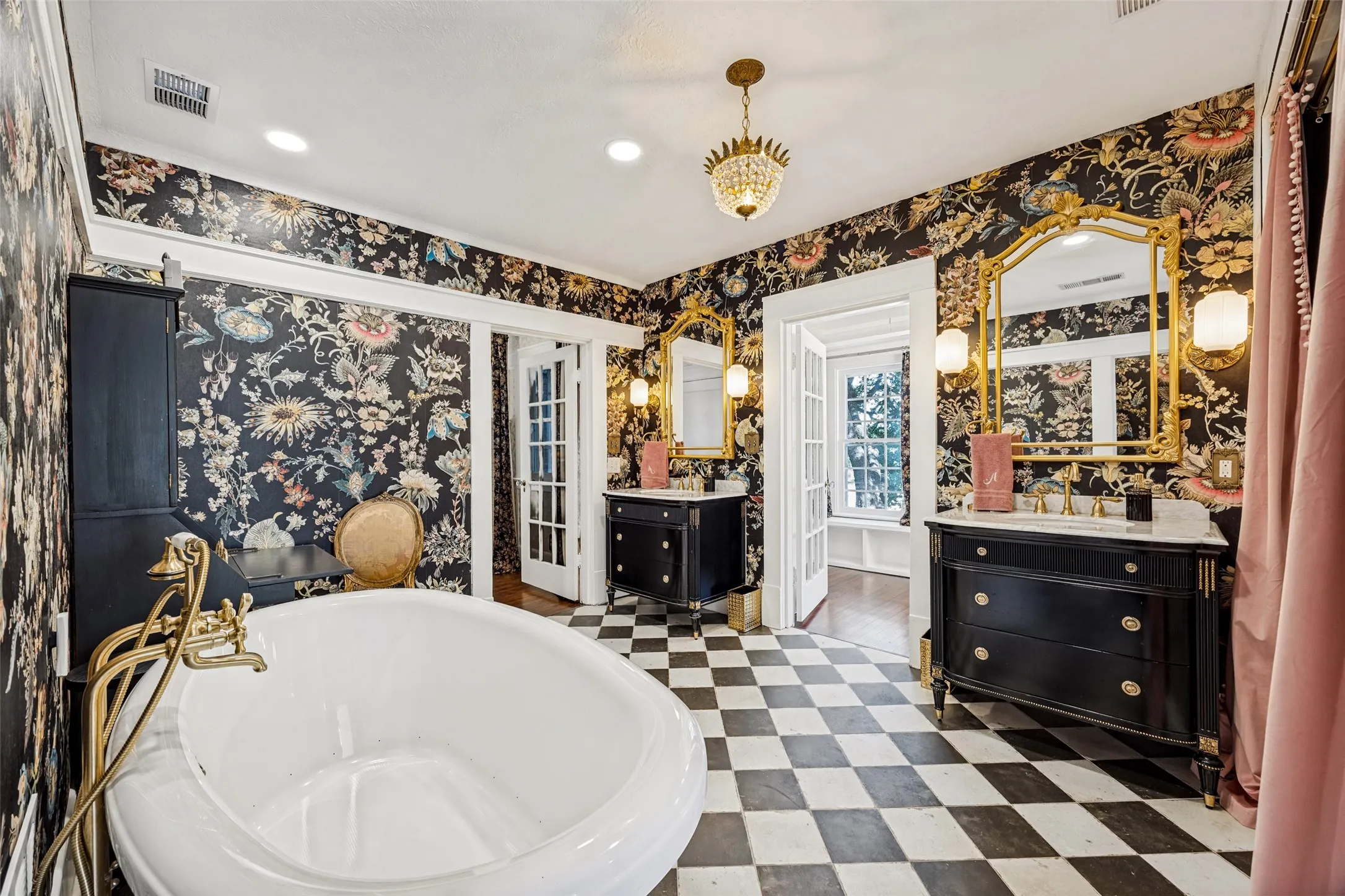 Freestanding soaking tub with a brass fixture, dual vanity sinks with marble tops, ornate gold-framed mirrors, and checkerboard floor tiling
